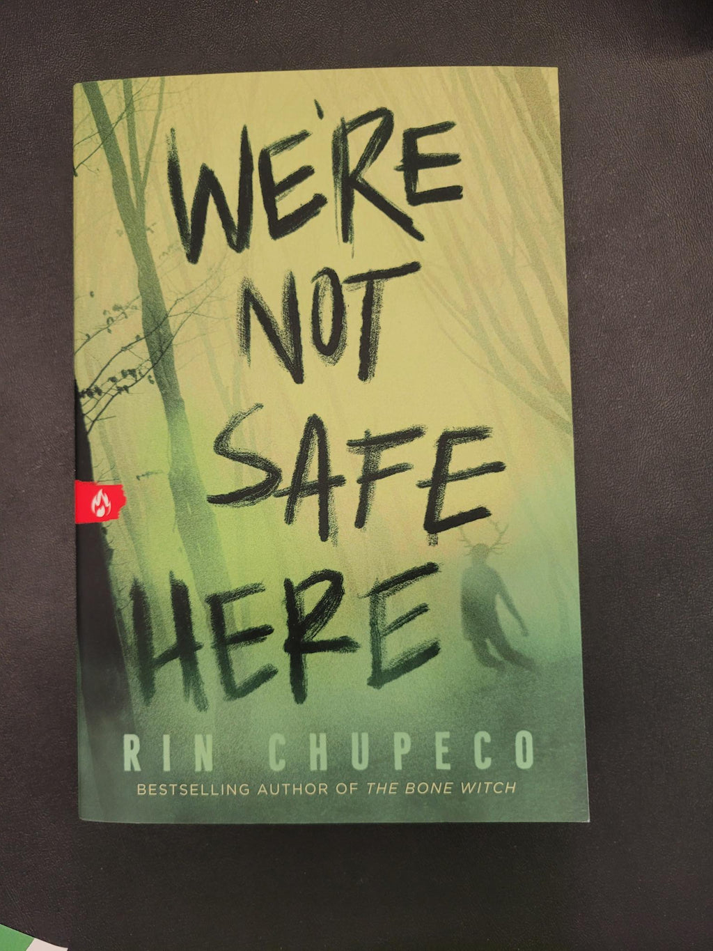 A book titled 'We're Not Safe Here' by Rinn Chupeco, featuring a green and yellow-green cover with black handwritten-style text, photographed on a dark surface.