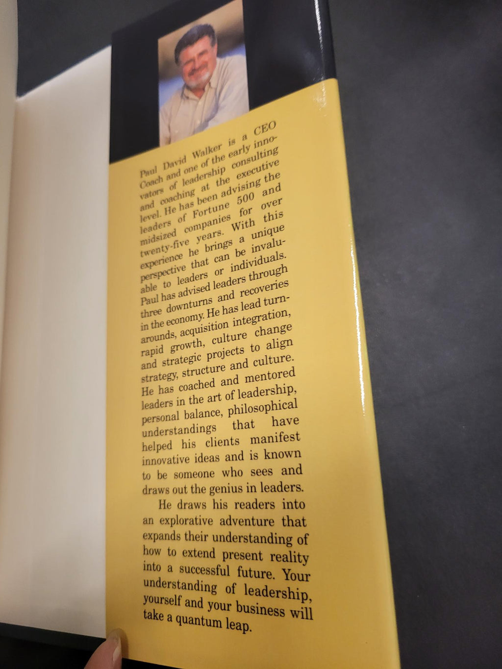 A close-up of a book or booklet cover with a yellow background, black side panels, and a small portrait photo of a man on the left. A hand is holding the item at the bottom right.