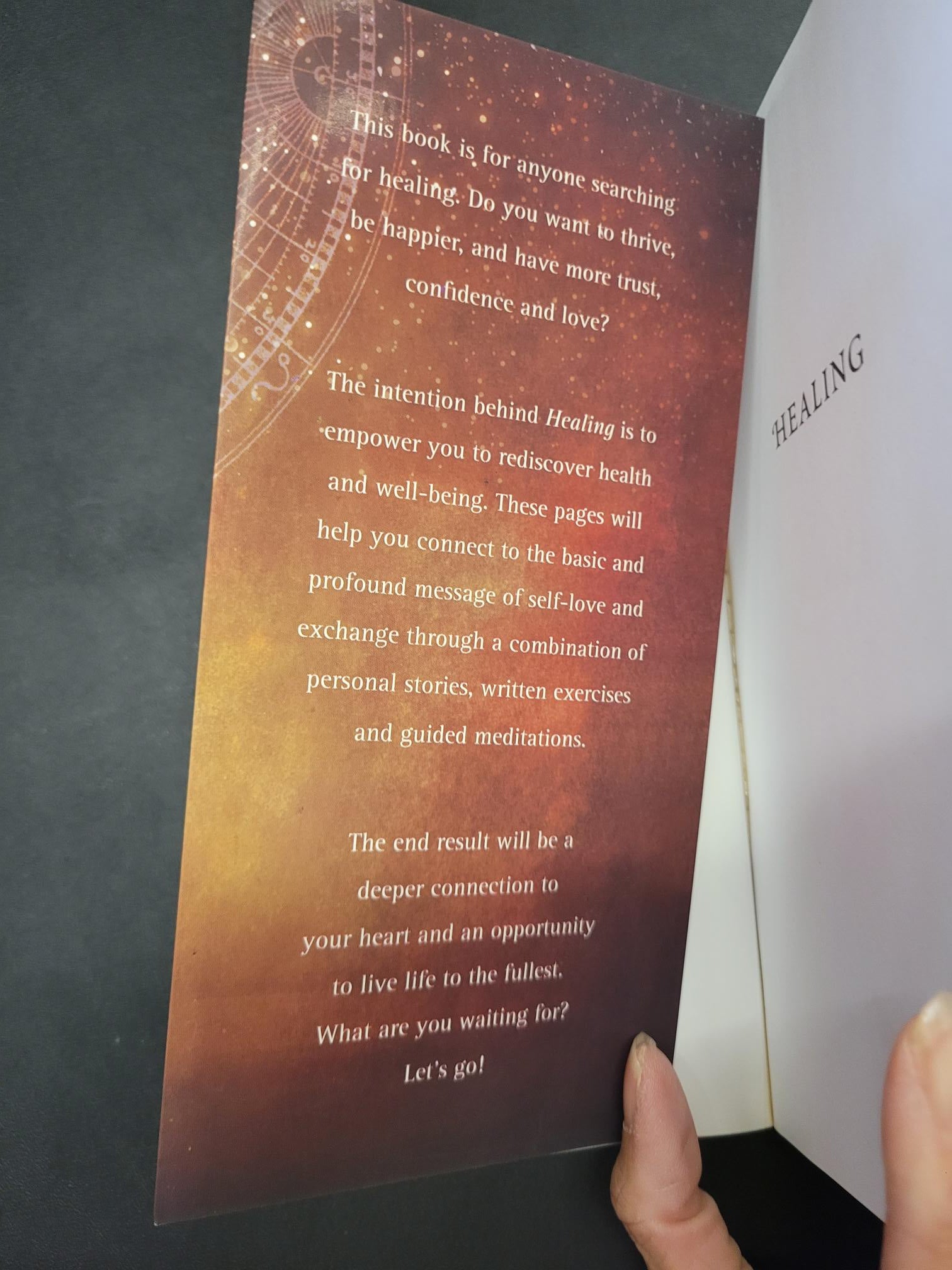 A person's hand holding a printed card or booklet page with inspirational text over a warm brown and gold gradient background, with part of a white book or box visible at the top.