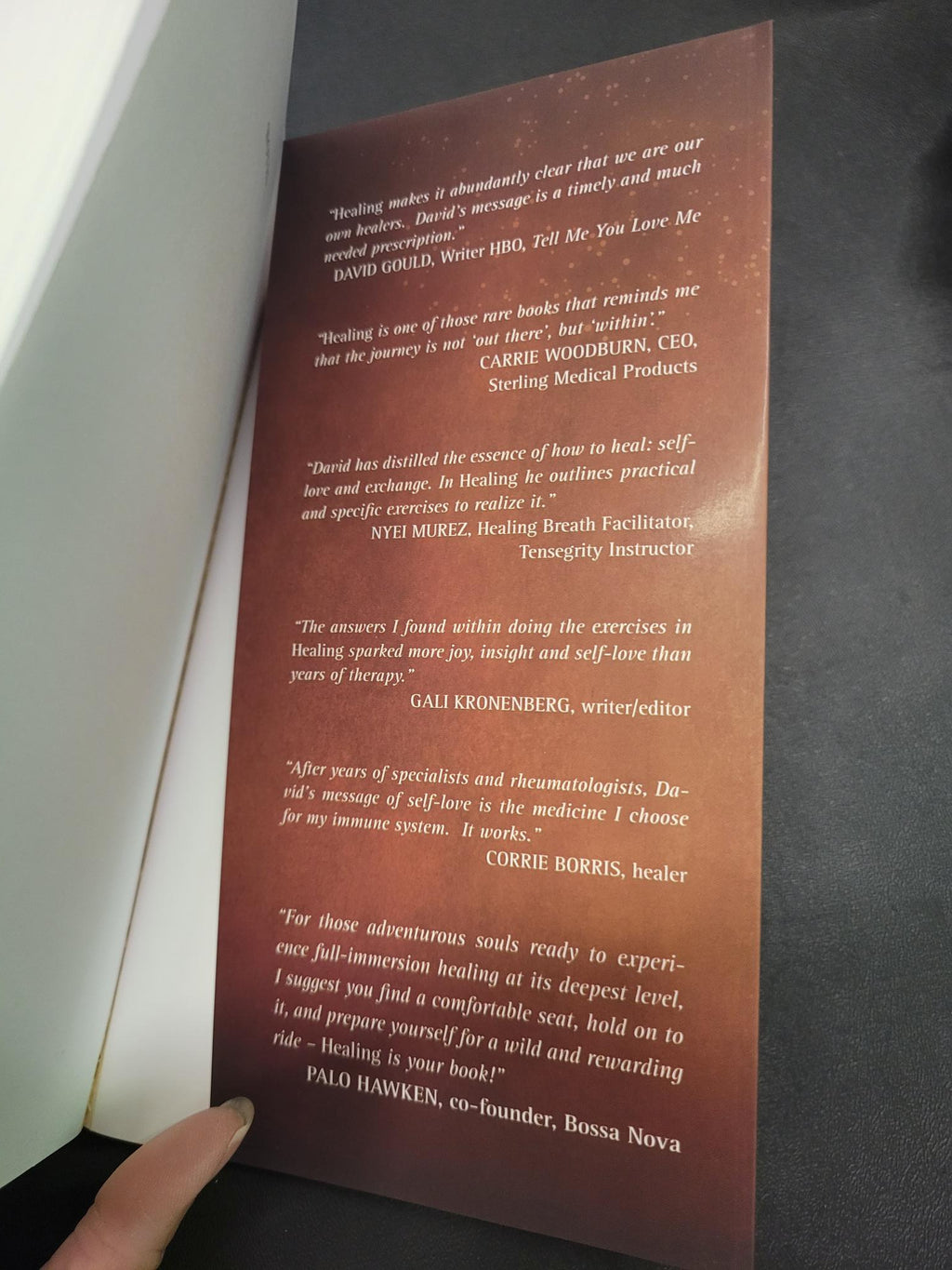 A hand holding open a book or booklet, showing a printed interior page with several paragraphs of text on a reddish-brown background.
