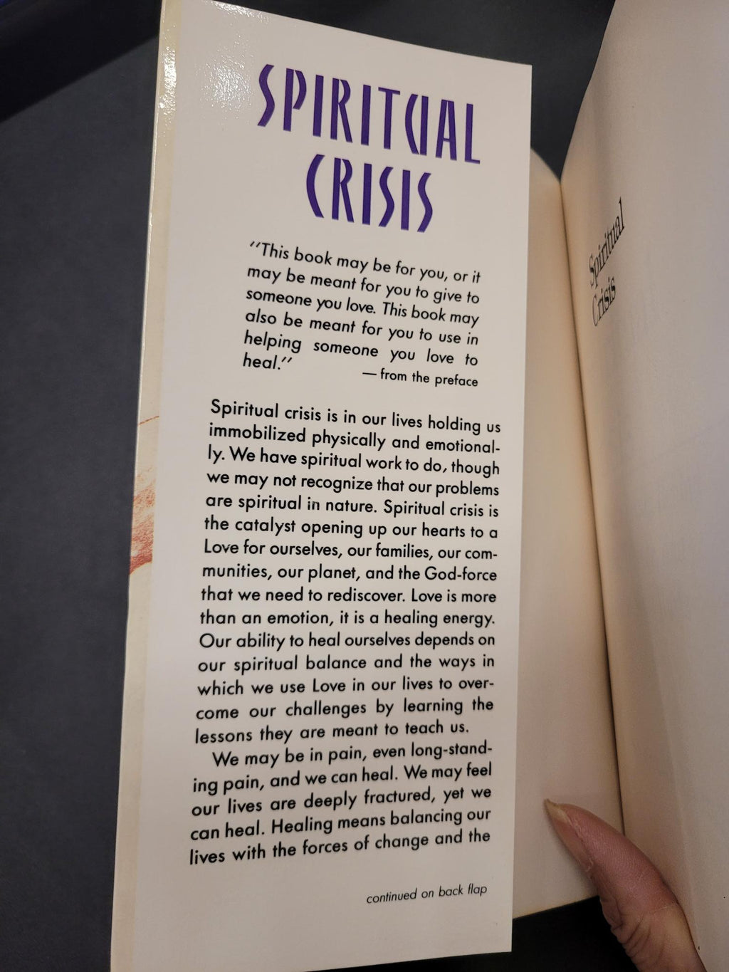 A close-up of a book's interior page titled 'SPIRITUAL CRISIS' with printed text, photographed inside an open book. A person's fingers are visible holding the page in place.
