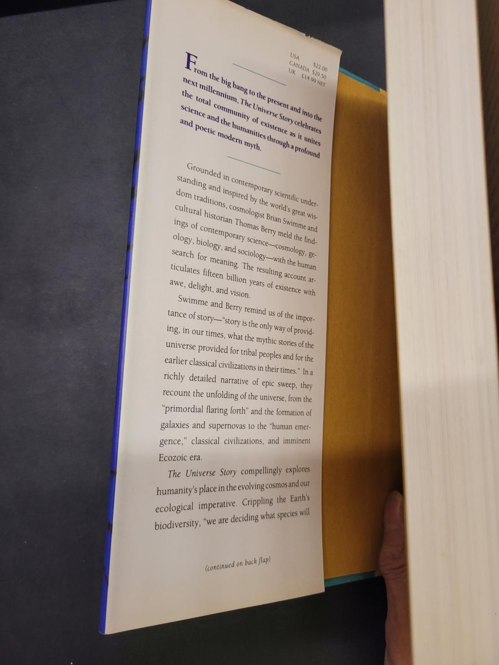 A printed page from a book or magazine is laid open on a dark surface, with a hand holding the top edge of the page or book. The page contains black text on off-white paper, and the book has a blue cover visible along the bottom edge.