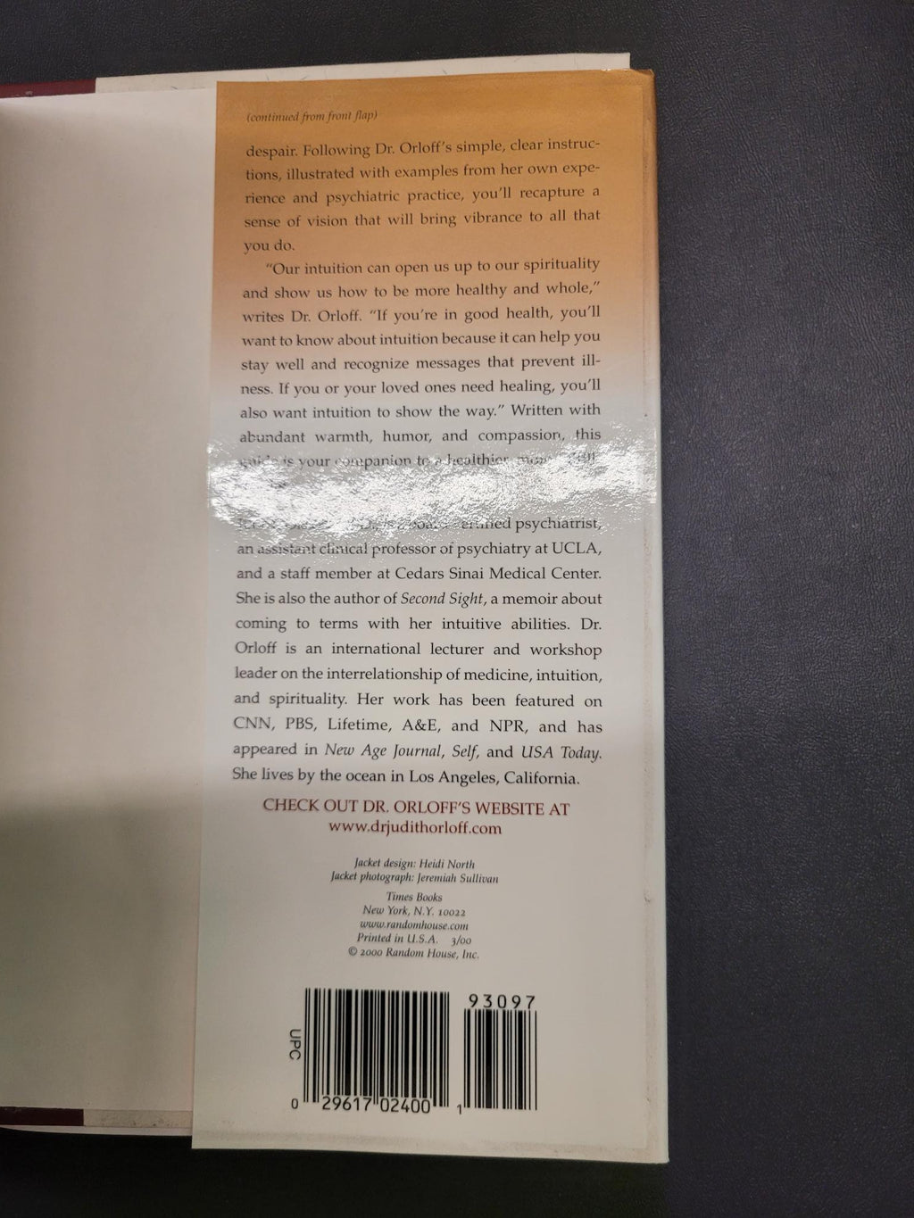 The back cover of a book lying on a dark gray surface, showing a tan and white design with text, a barcode, and a clear protective sticker in the center.
