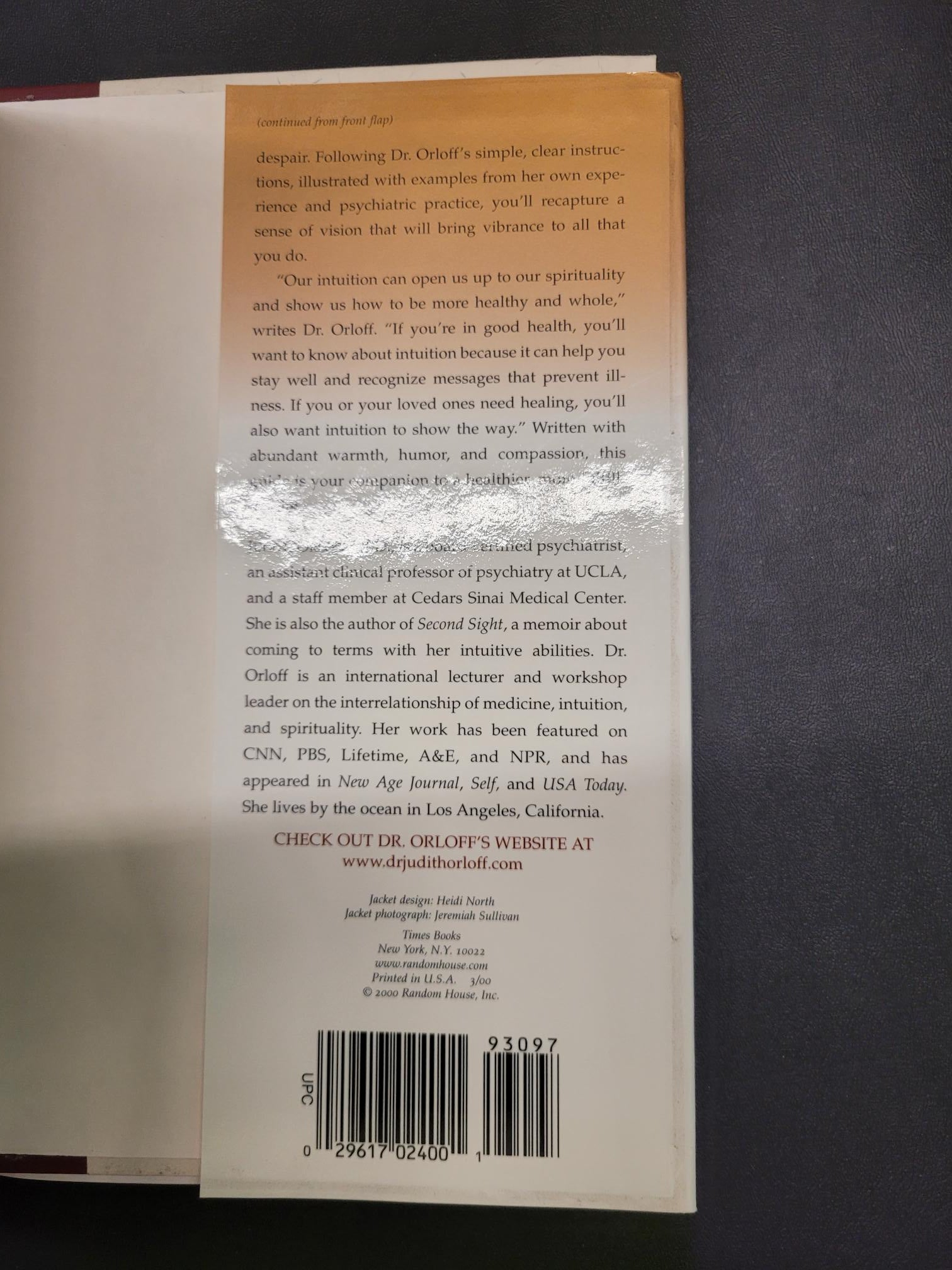 The back cover of a book lying on a dark gray surface, showing a tan and white design with text, a barcode, and a clear protective sticker in the center.