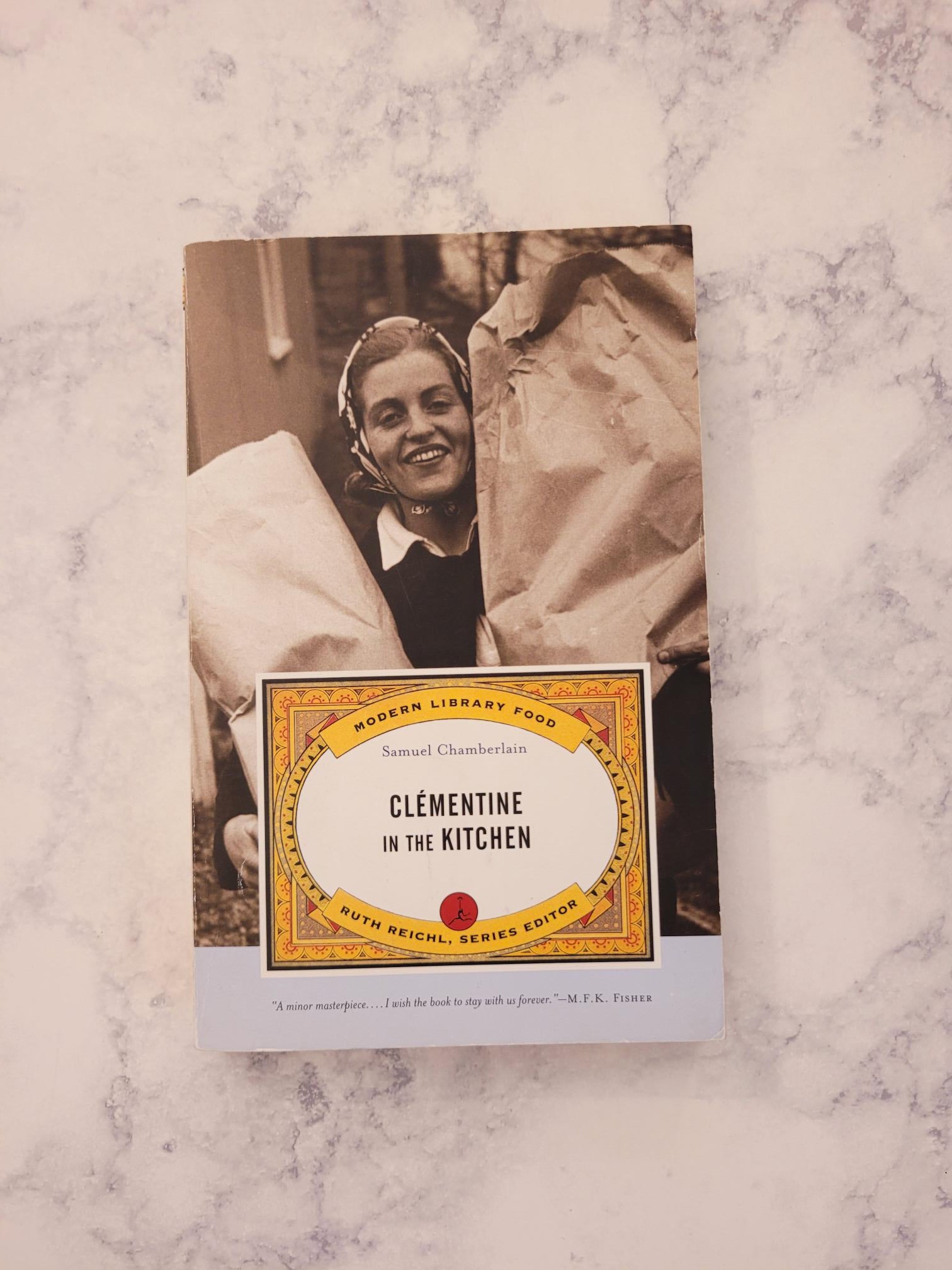 A book titled 'Clementine in the Kitchen' by Samuel Chamberlain, featuring a vintage-style cover with a sepia-toned photograph of a woman and a gold-bordered title label, placed on a light marble surface.