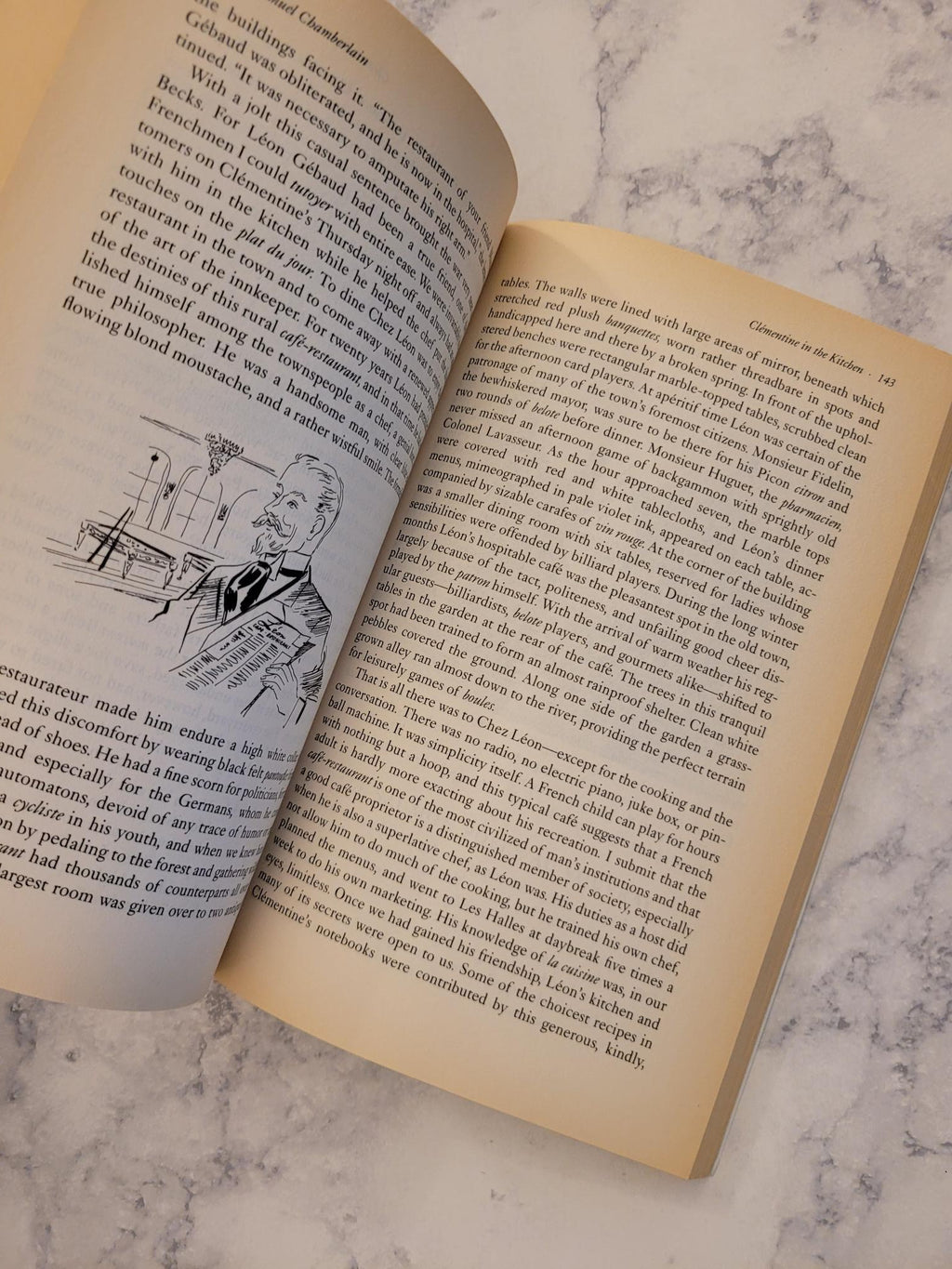 An open vintage book with aged cream-colored pages, displaying dense black text and a small black-and-white illustration on one page, set against a light gray marble background.