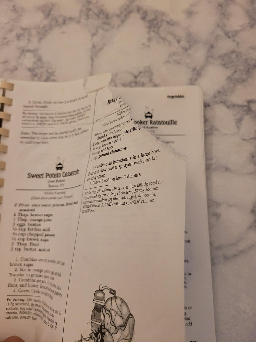 A spiral-bound recipe booklet or cookbook page showing printed text and a small illustration, photographed on a white marble surface.