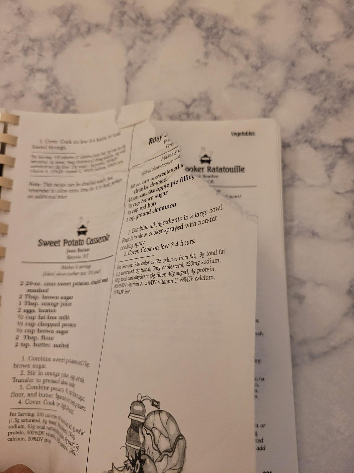 A spiral-bound recipe booklet or cookbook page showing printed text and a small illustration, photographed on a white marble surface.