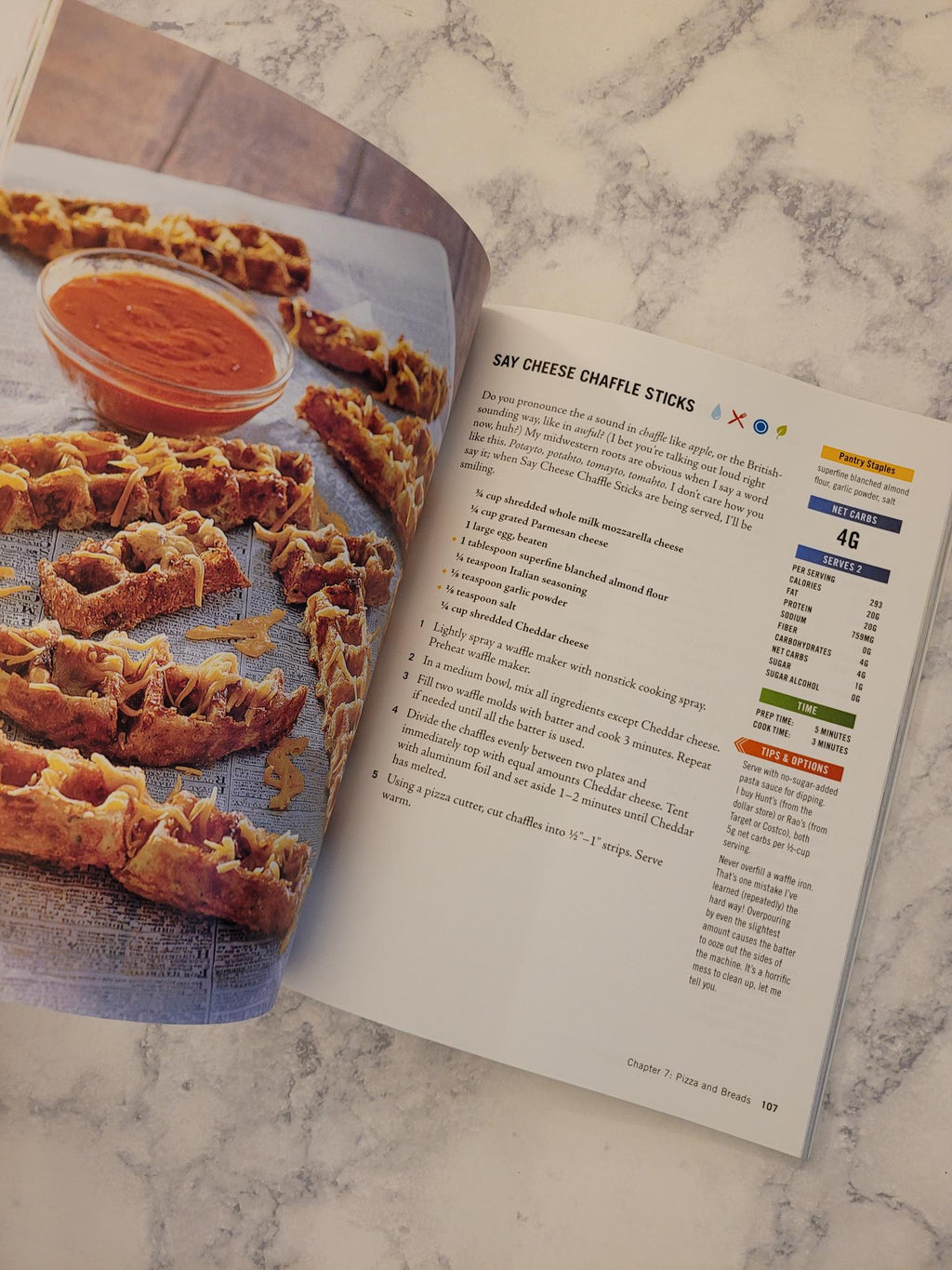 An open cookbook spread on a light marble surface, showing the recipe page for sauté cheese dumpling sticks with a printed photo of the finished stuffed dumplings and a small bowl of orange sauce.