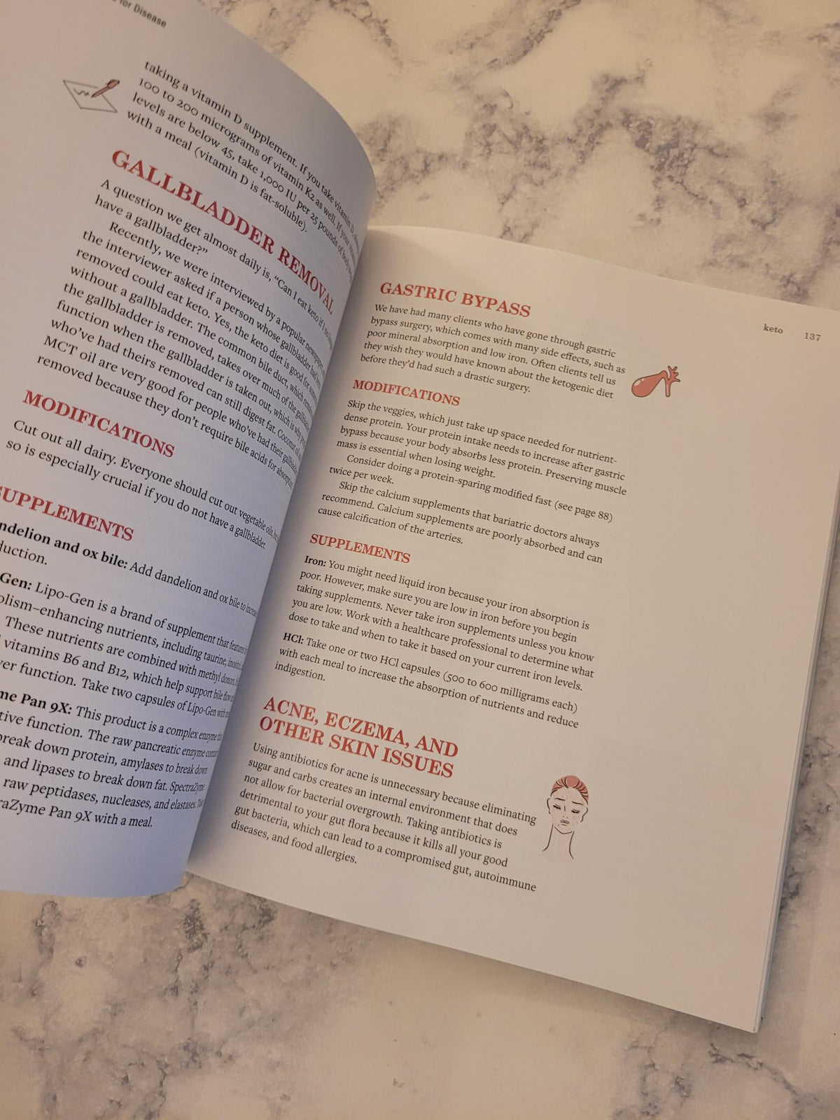 An open printed booklet or recipe/guide book laid on a light marble surface, showing printed text columns with small red and black illustrations. No person or animal is present, and the scene is a styled flat-lay rather than a real environment emphasis, so this fits Product/Packaging rather than Lifestyle.