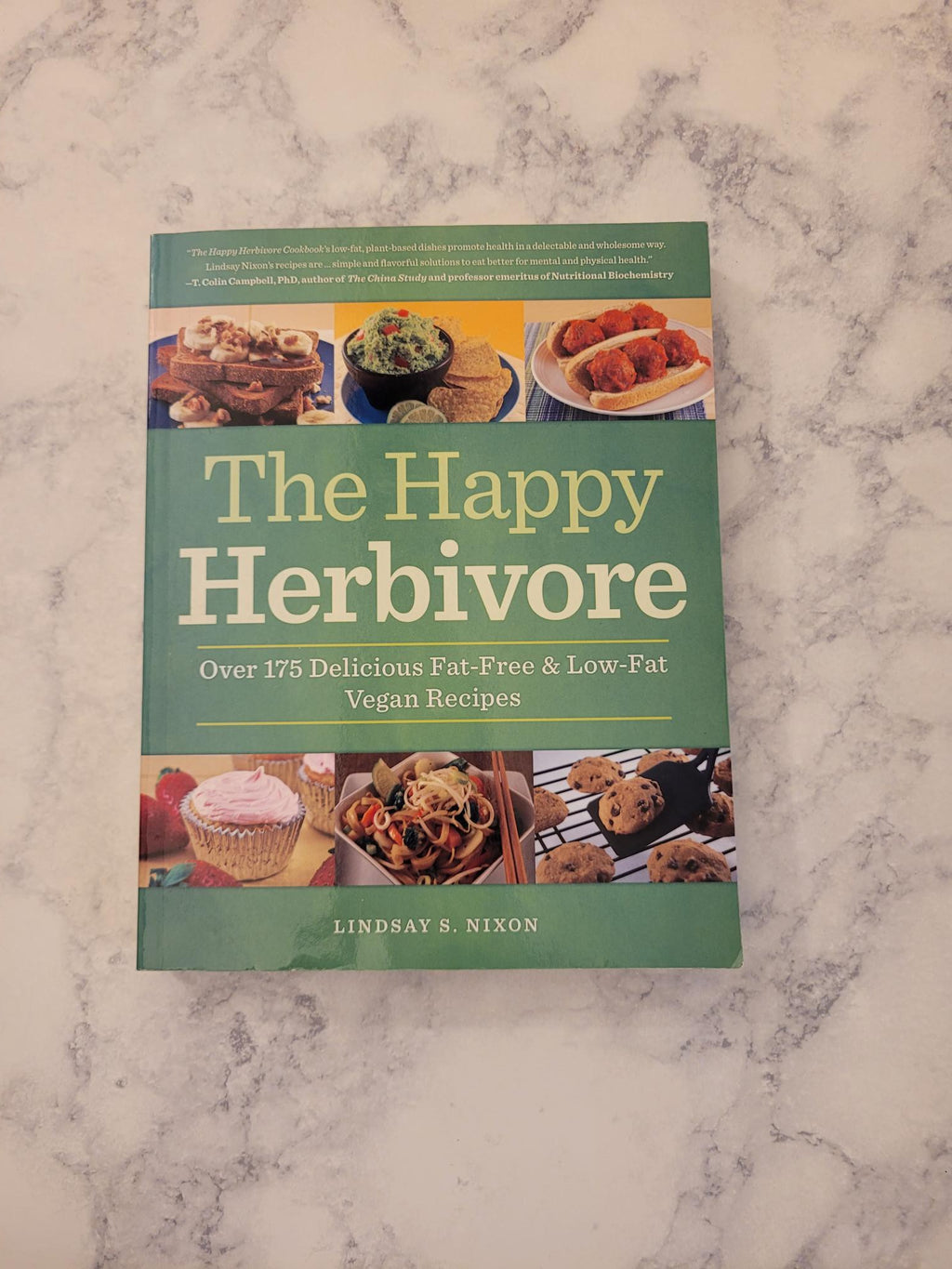 The Happy Herbivore cookbook by Lindsay S. Nixon, featuring a sage green cover with food photography, displayed on a white and gray marble surface.