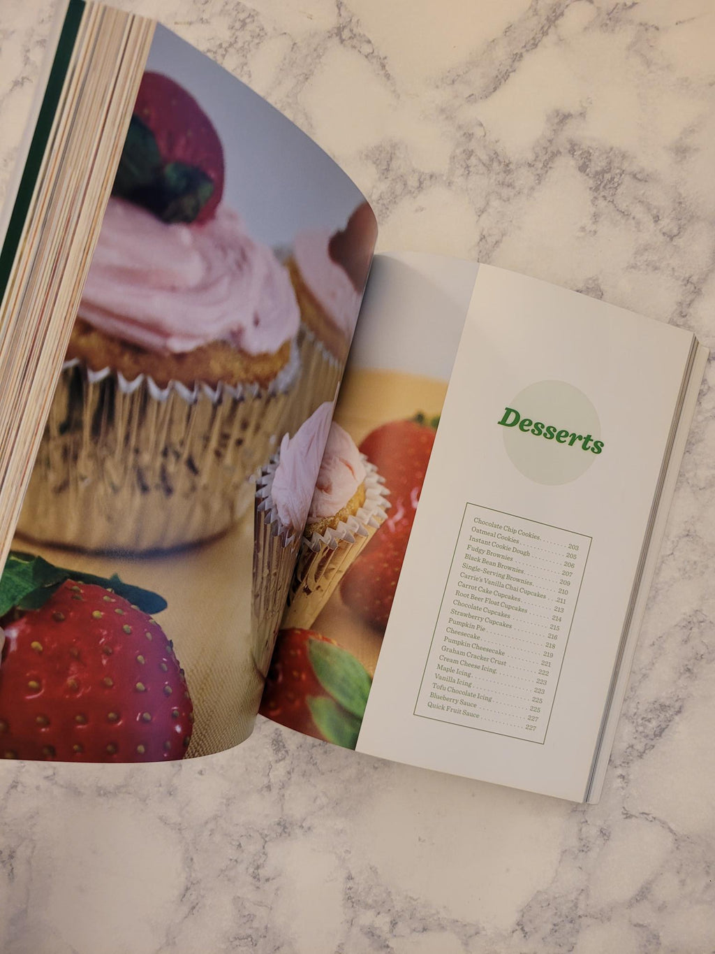 An open cookbook spread showing a dessert page with a cupcake photo and a printed index, placed on a white marble surface.