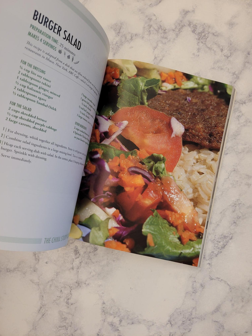 An open cookbook or recipe book lying on a light marble surface, with a colorful photo of a burger salad on the top page and the recipe text for 'Burger Salad' visible on the bottom page.