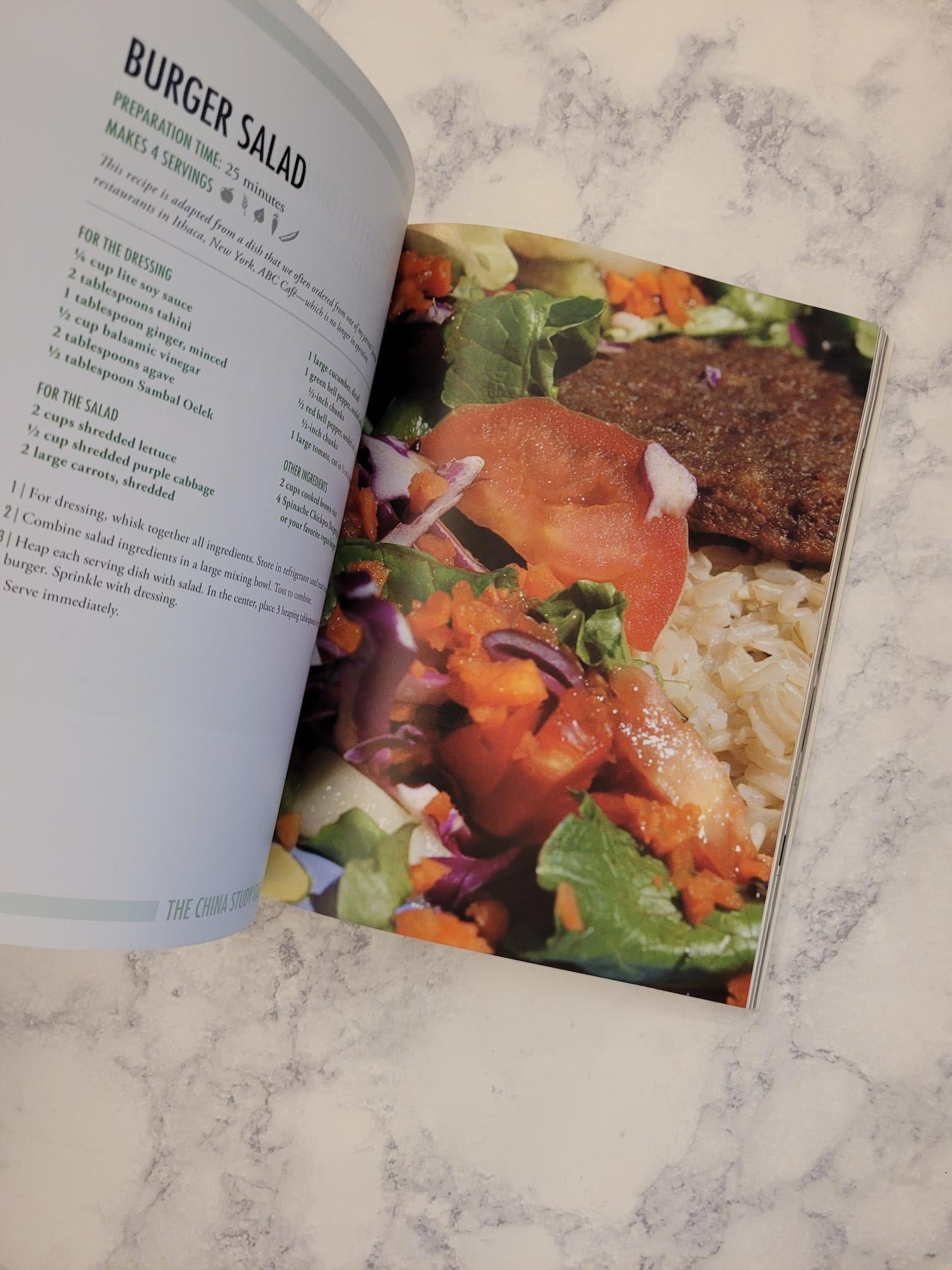 An open cookbook or recipe book lying on a light marble surface, with a colorful photo of a burger salad on the top page and the recipe text for 'Burger Salad' visible on the bottom page.