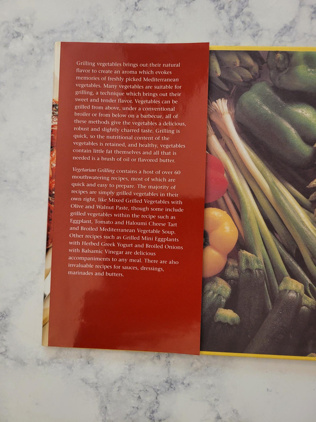 A printed booklet or book page showing a close-up photo of assorted vegetables on the top half and a large red text block on the bottom half, photographed on a light marble-like surface.