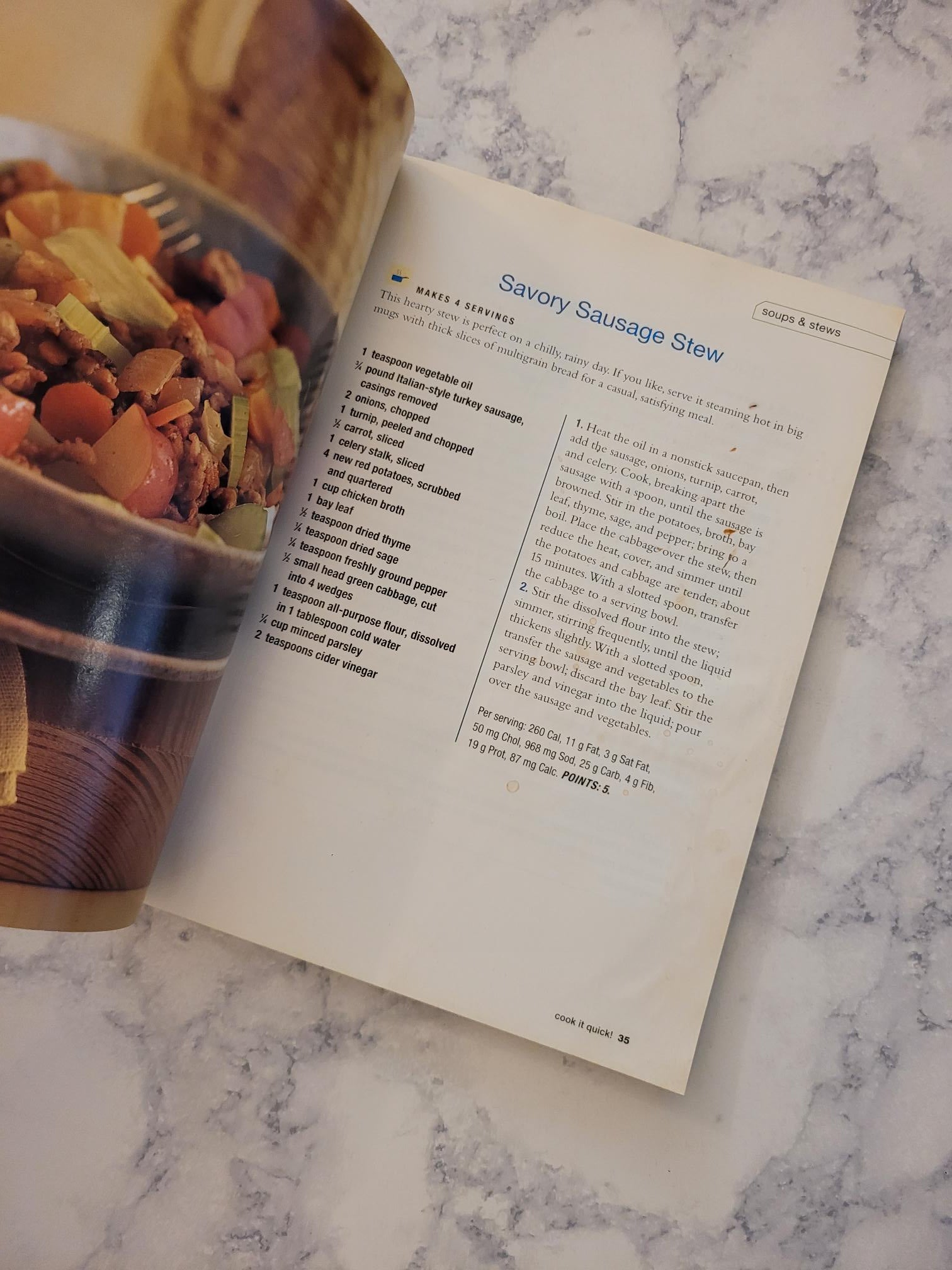 An open cookbook or recipe book lying on a white marble surface, showing a page titled 'Savory Sausage Stew' with text and a photo of a stew dish at the bottom.