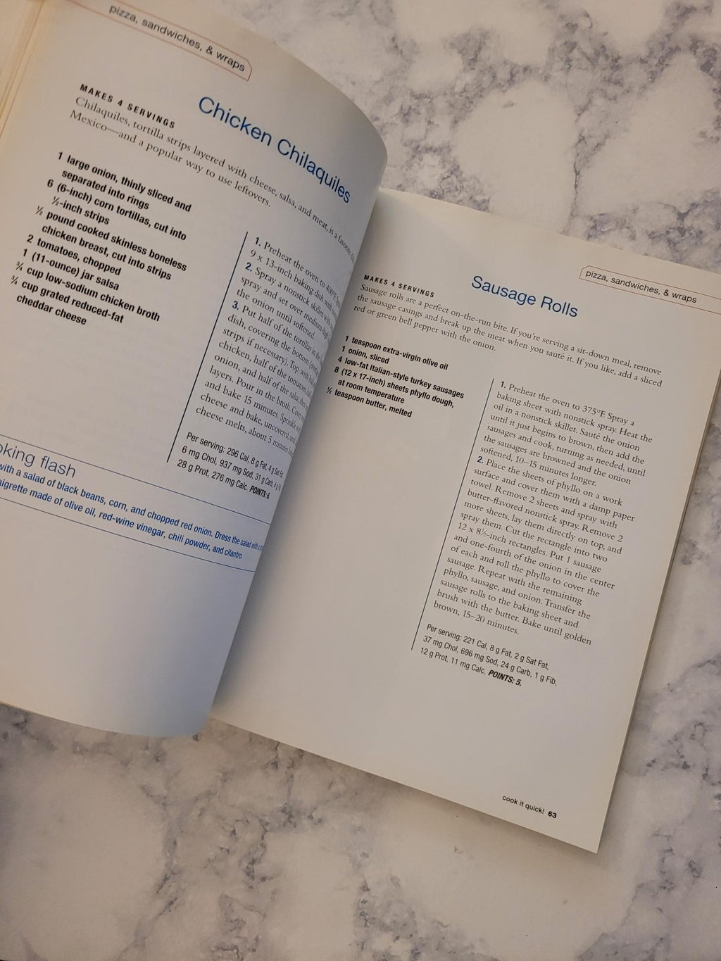 An open cookbook or recipe book spread on a white and gray marble surface, showing printed recipe pages with headings like 'Sausage Rolls' and 'Chicken Chilaquiles'.