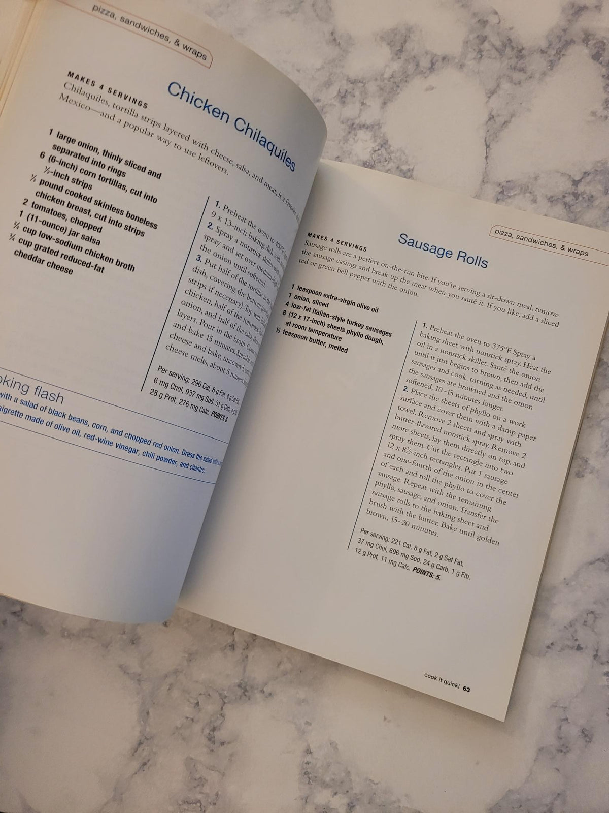 An open cookbook or recipe book spread on a white and gray marble surface, showing printed recipe pages with headings like 'Sausage Rolls' and 'Chicken Chilaquiles'.