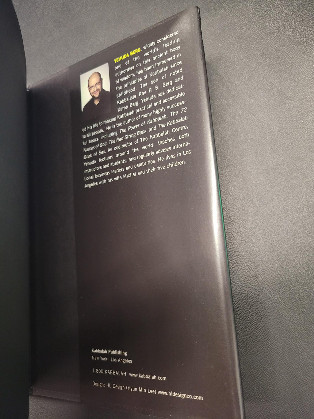 The back cover of a book or booklet lying on a dark gray textured surface. The cover is dark brown with white text, a small portrait photo of a man, and contact information printed on the right side.