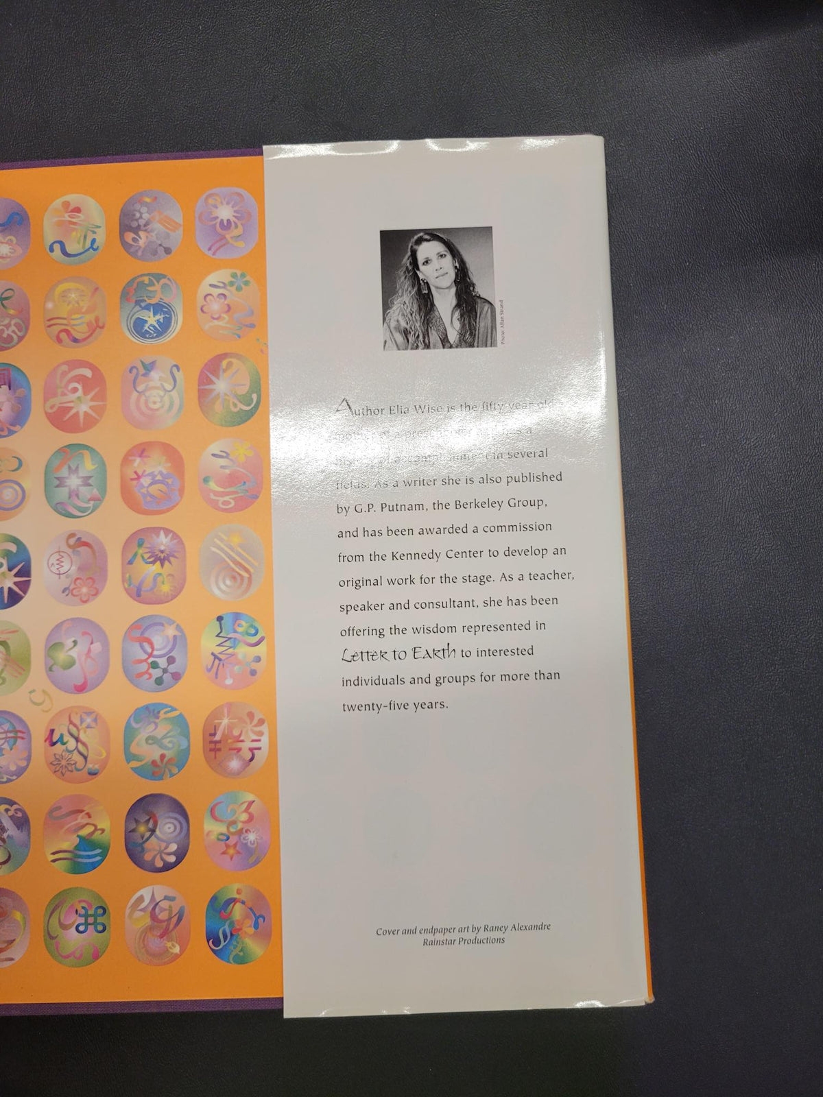 An open book or booklet lying on a dark surface, showing the back cover with a black-and-white portrait photo, small text, and a row of colorful circular illustrations along the bottom.