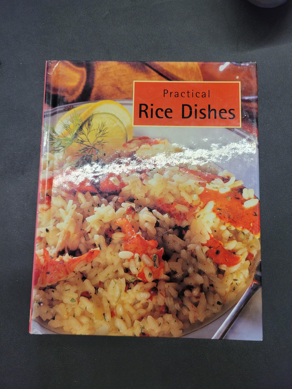 A book titled 'Practical Rice Dishes' lying on a dark gray surface. The cover features a close-up photograph of a rice dish with orange-red vegetables and a lemon garnish.