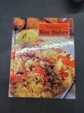 A book titled 'Practical Rice Dishes' lying on a dark gray surface. The cover features a close-up photograph of a rice dish with orange-red vegetables and a lemon garnish.