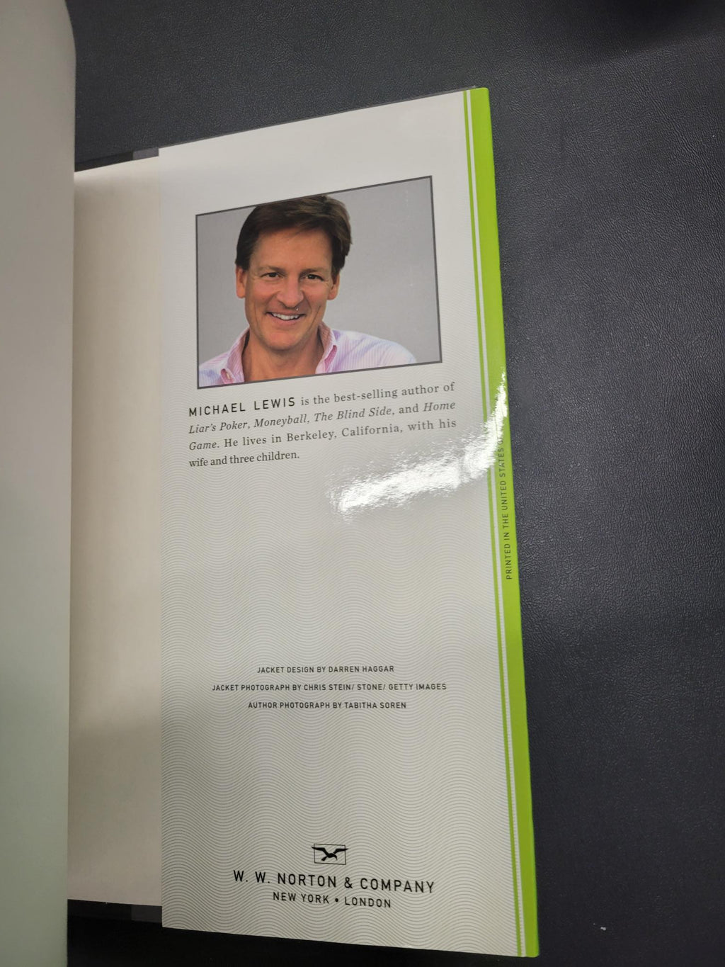 A printed booklet or brochure page shown at an angle on a dark surface. The page has a white background with a bright green horizontal band near the top, a small portrait photo of a smiling man, biographical text, and the visible brand/title 'W.