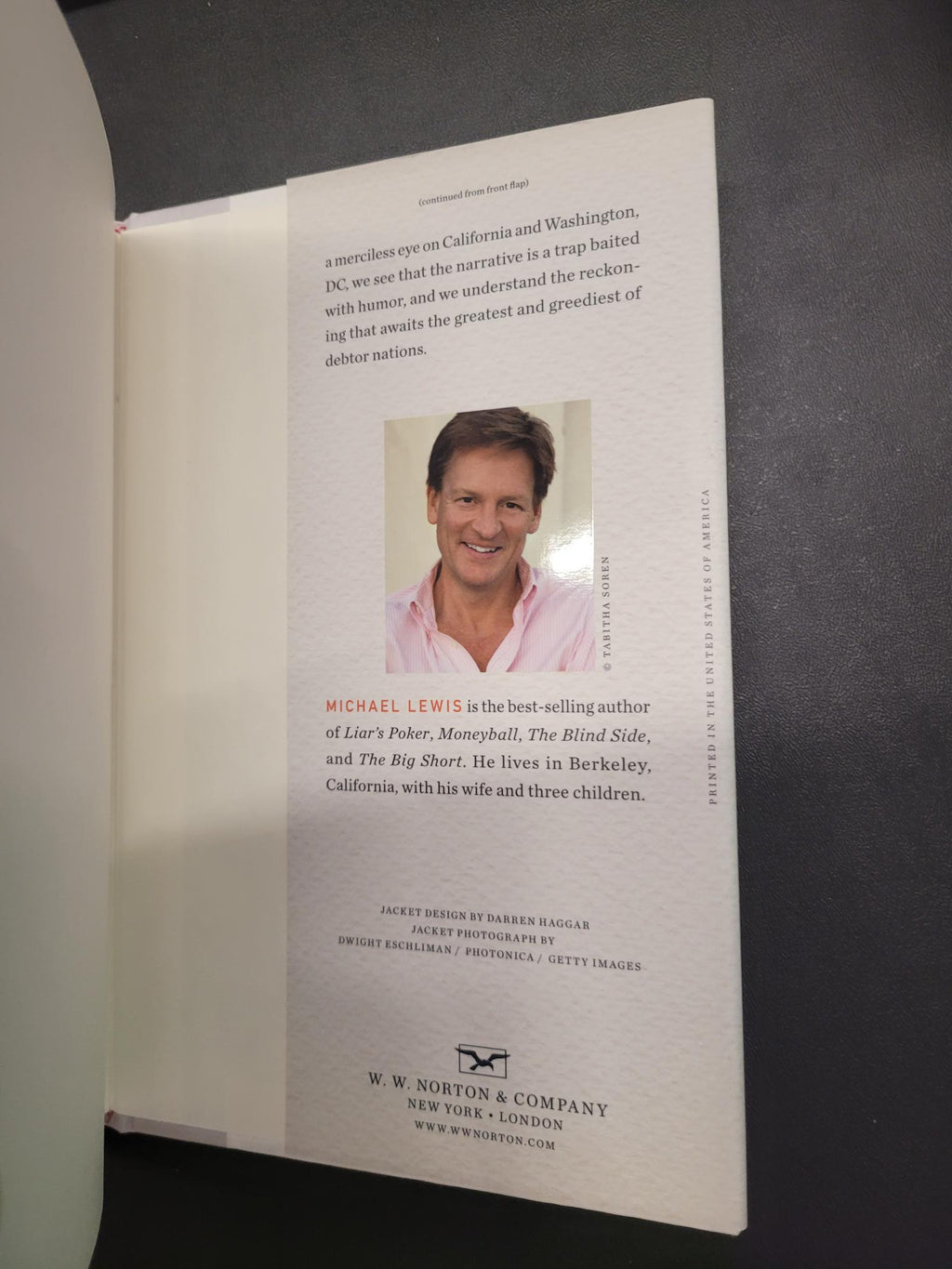 An open book or booklet showing printed text and a small portrait photo of Michael Lewis on a light page, photographed against a dark background.