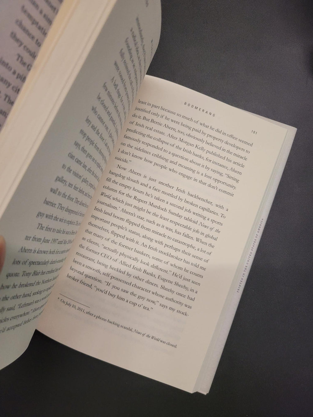 An open book showing printed text on two pages, photographed against a dark gray surface. A finger is visible holding the lower page.