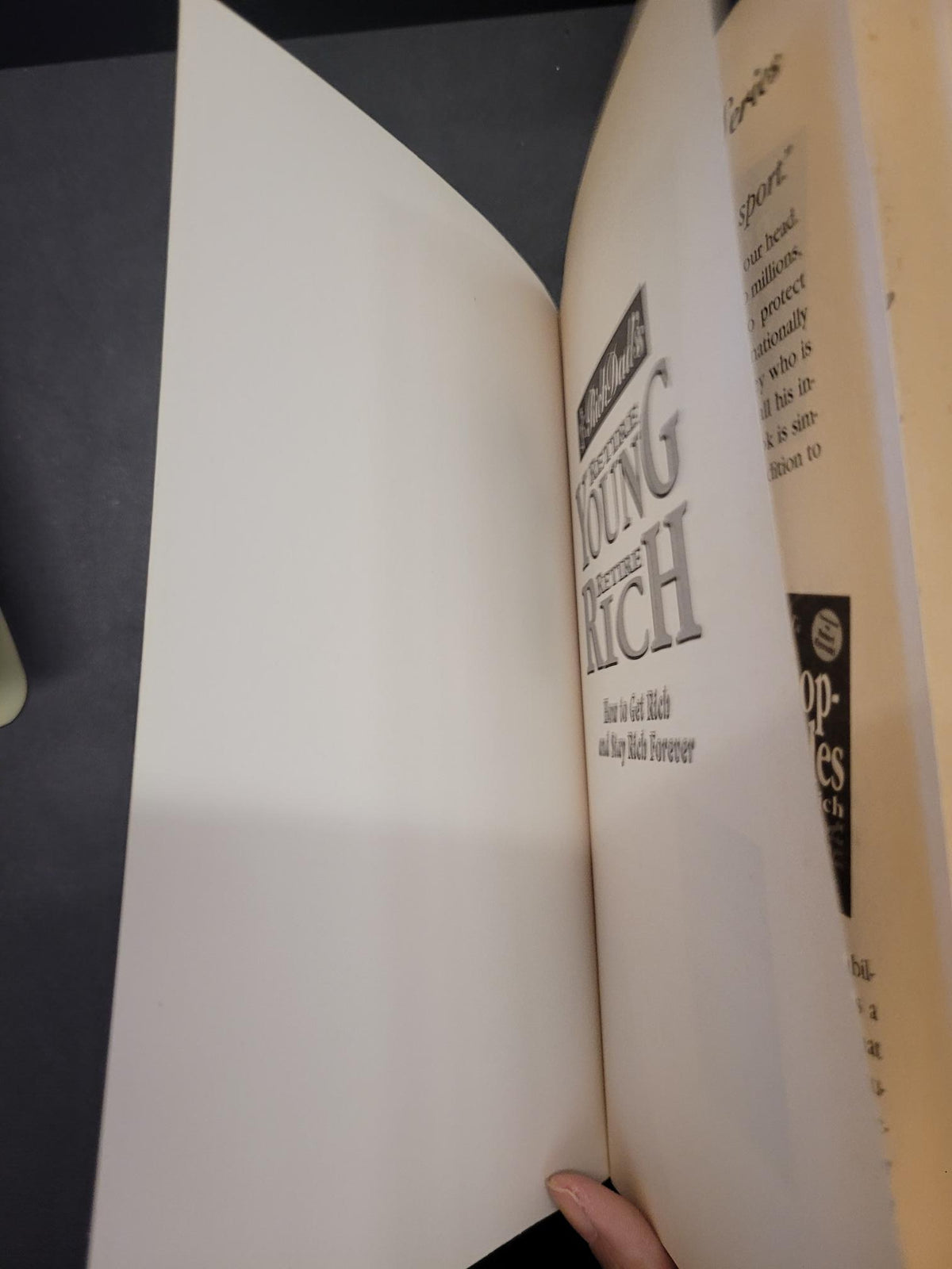 A hand holding open a large white book or binder, showing printed text and pages against a dark tabletop background.