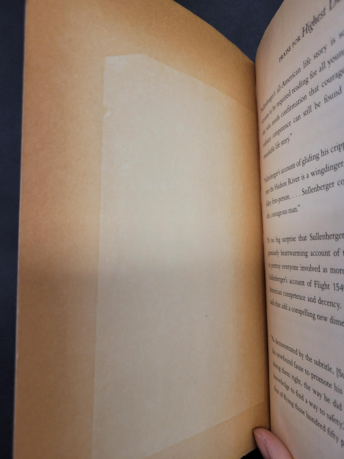 A close-up of an open book showing blank, unprinted tan paper sheets with a large rectangular translucent protective cover over one page. A finger is visible at the top right holding the page open.