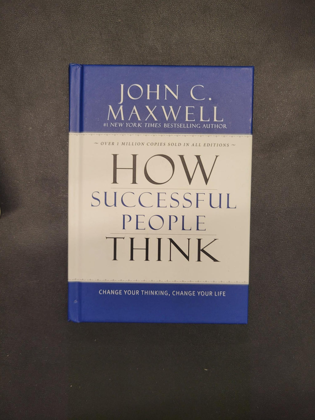 The book 'How Successful People Think' by John C. Maxwell, featuring a blue and white cover, photographed on a dark gray textured surface.
