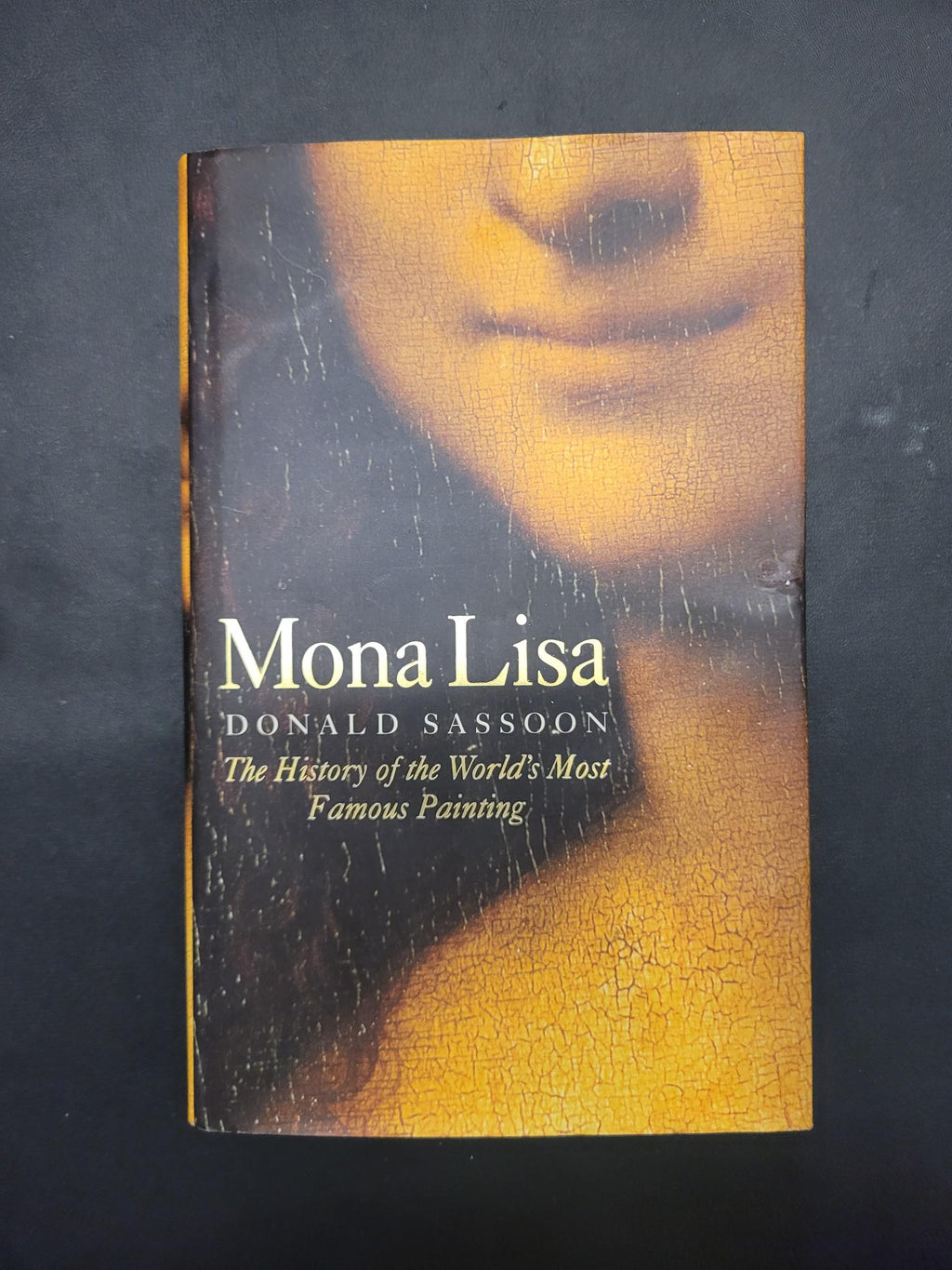 The book cover of 'Mona Lisa' by Donald Sassodon, featuring a close-up of Leonardo da Vinci's painting with a cracked yellow-gold finish and white text, photographed on a dark textured background.