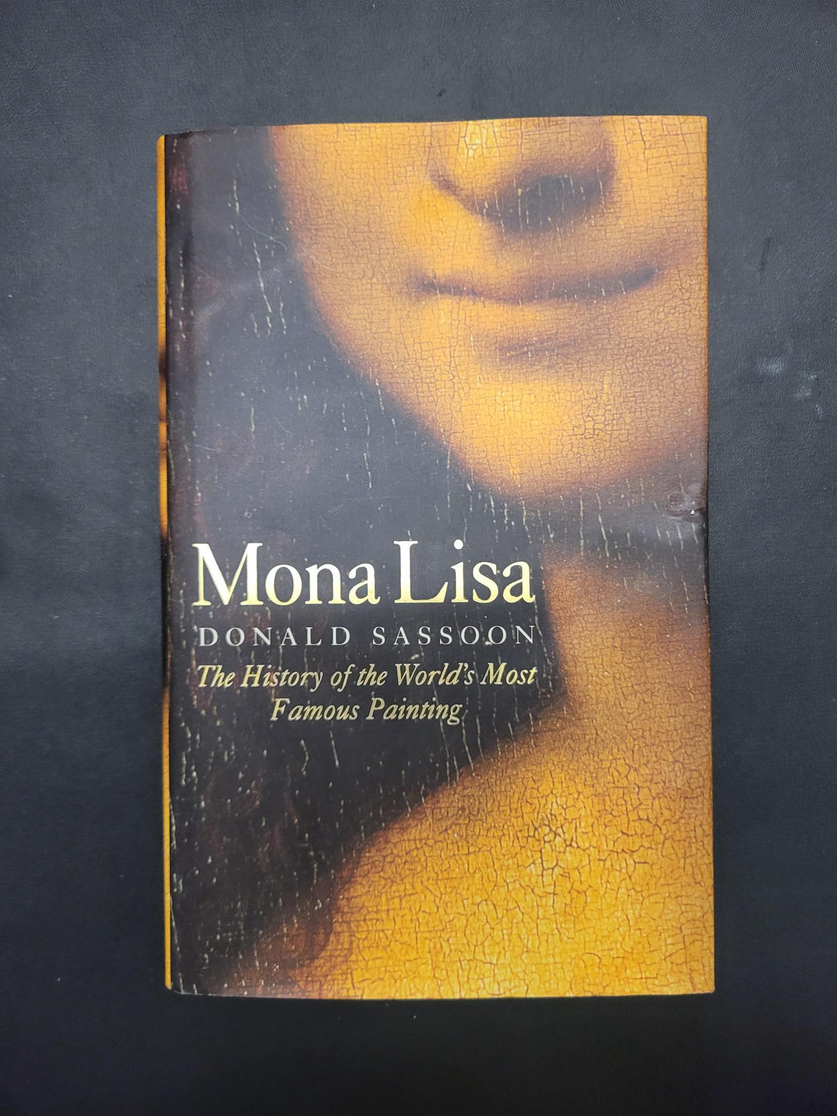 The book cover of 'Mona Lisa' by Donald Sassodon, featuring a close-up of Leonardo da Vinci's painting with a cracked yellow-gold finish and white text, photographed on a dark textured background.