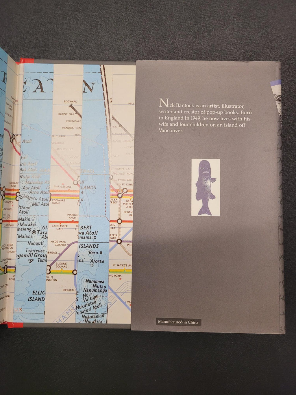 A book or booklet shown open on a dark surface, with the top page in gray featuring a small fish illustration and text, and the lower pages showing a map with blue water areas and multicolored route lines.