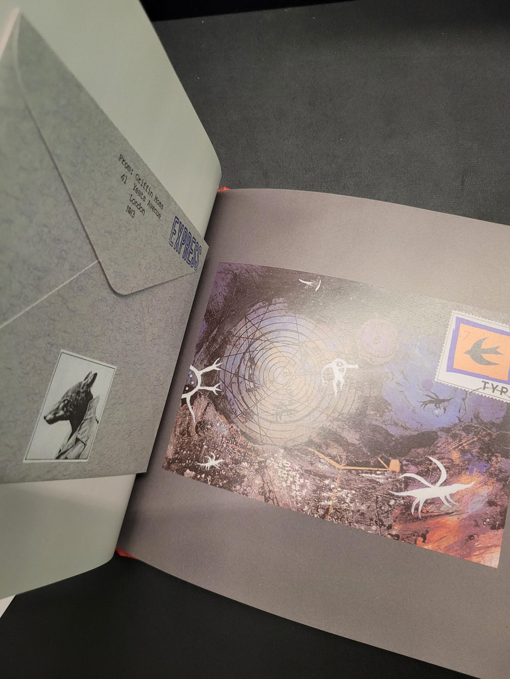 A gray textured envelope with a printed address and a small black-and-white photo insert, placed in front of an open book or booklet showing a colorful abstract cover illustration.