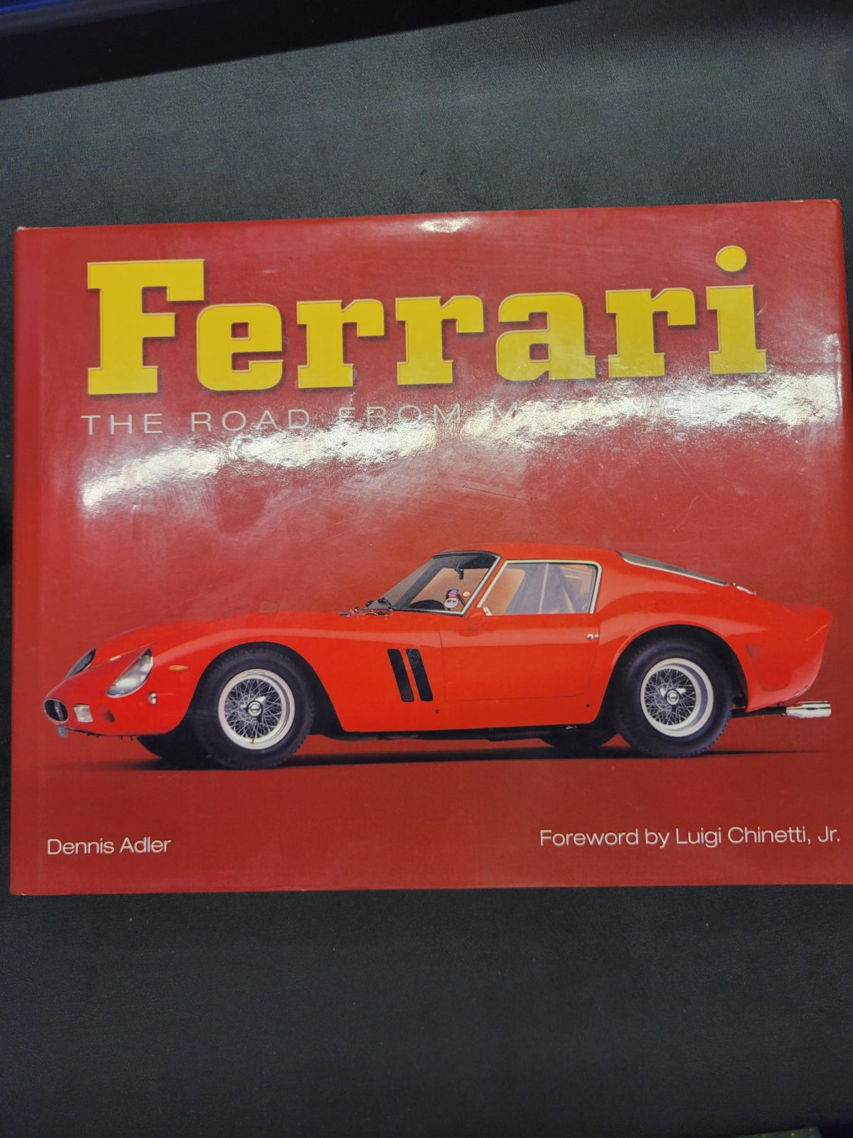 A book titled "Ferrari: The Road From War" with a bright red cover, large yellow Ferrari logo, and a red sports car illustration, photographed on a dark surface.