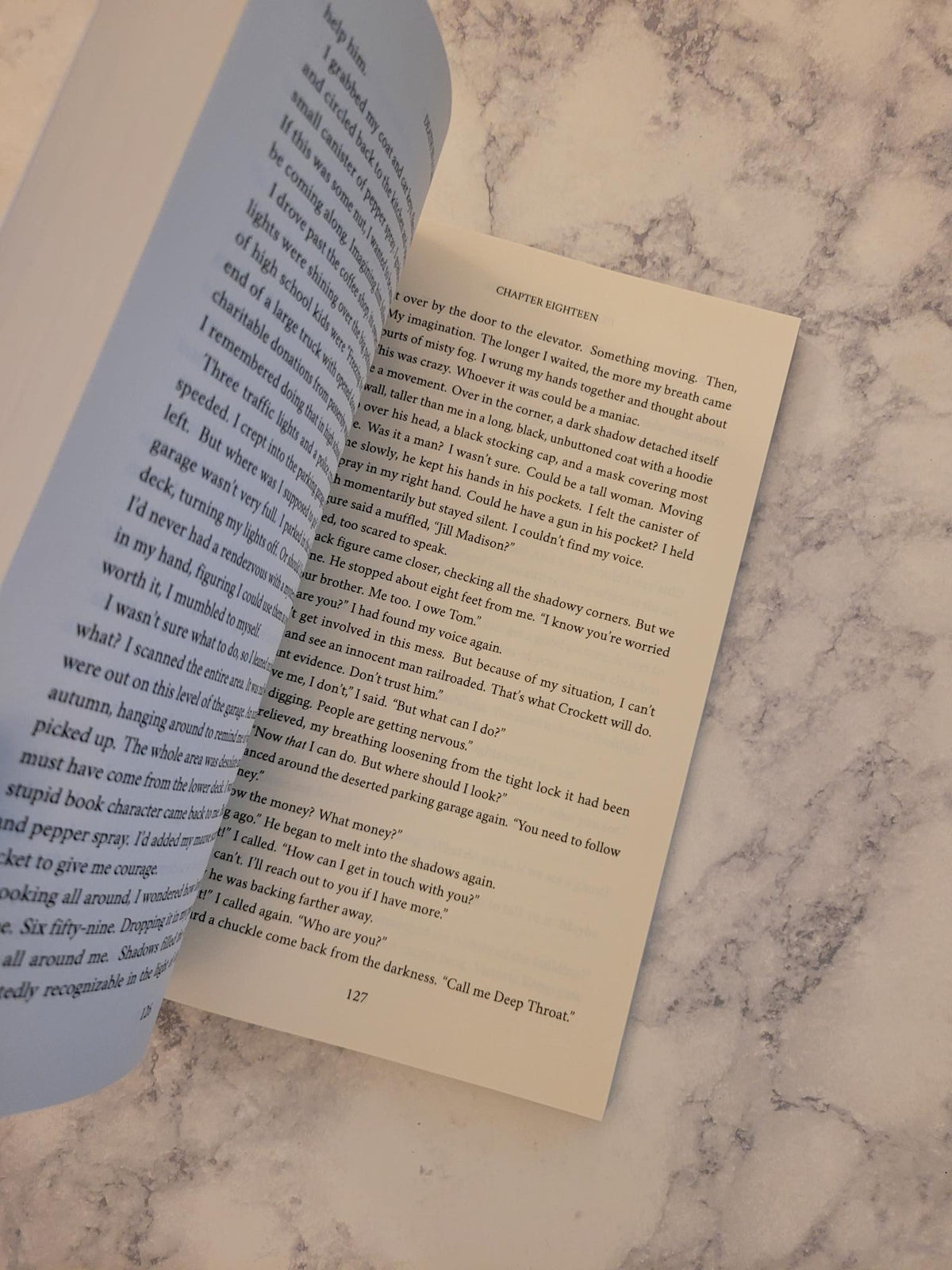 An open book lying on a white marble surface, showing printed text pages with a light blue left page and cream-colored right page.