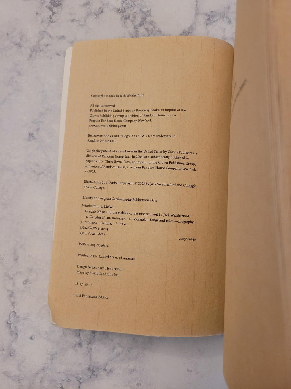An open book or booklet lying on a light marble-like surface, showing printed text on tan paper pages. A tan cylindrical object is partially visible along the top edge.