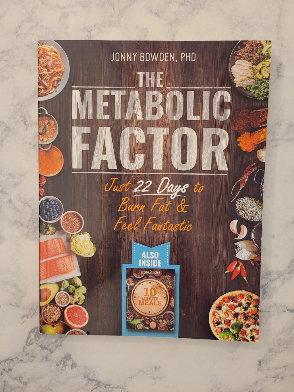The book cover of 'The Metabolic Factor' by Jonny Bowden, PhD, displayed flat on a light gray marble surface. The cover features a dark wood background with various food illustrations around the edges and a blue 'Also Inside' 10 Meal Plan booklet shown at the bottom.