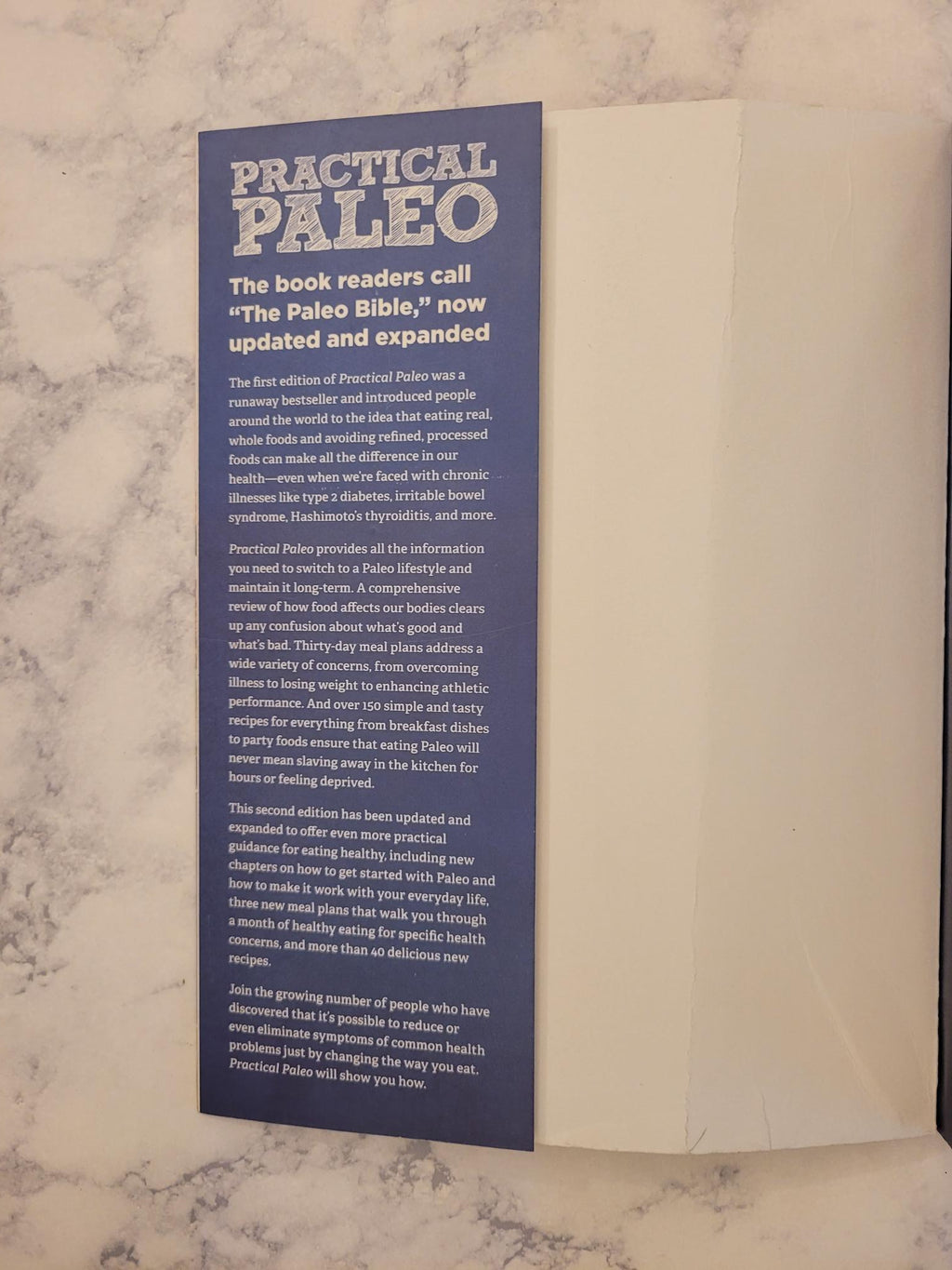 A printed insert or booklet page from the Practical Paleo book, featuring a dark blue background with white text and a large title, placed on a light marble-like surface next to a white envelope.