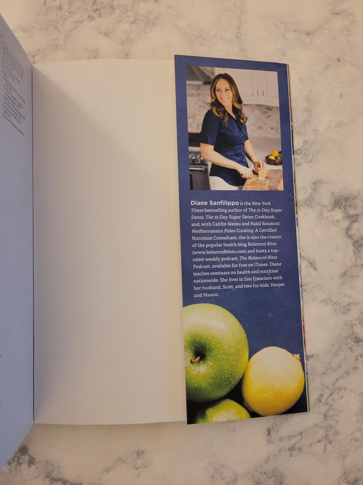 An open printed booklet or recipe book on a light marble surface, showing a printed page with a photo of a woman, text, and images of green apples and a yellow lemon on a dark blue background.