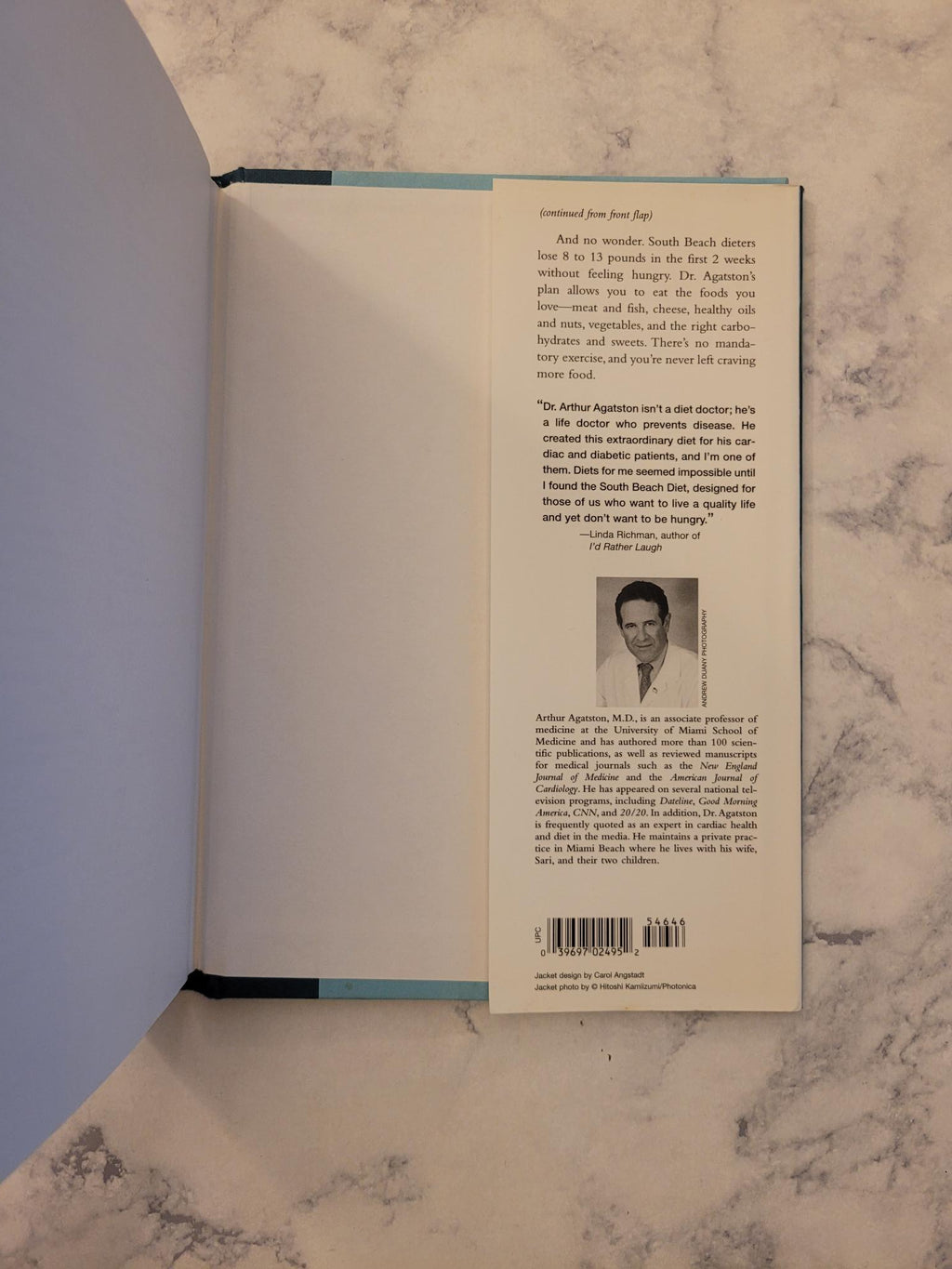 An open book showing the back cover, photographed from above on a light marble surface. The pages are spread open, with printed text and a small black-and-white portrait visible on the upper page.