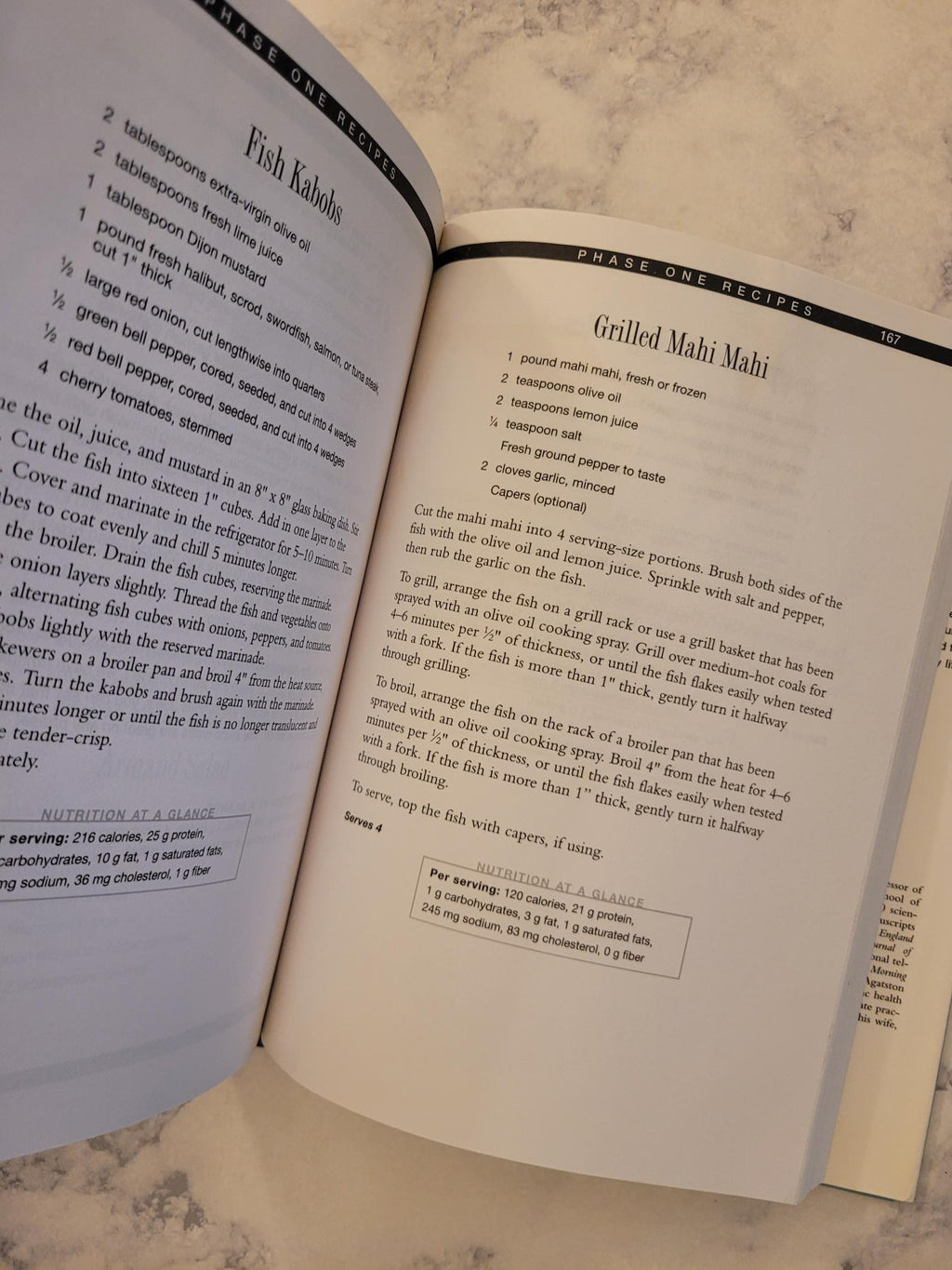 An open cookbook or recipe book spread on a light marble-like surface, showing printed recipe pages with headings and paragraphs of text.