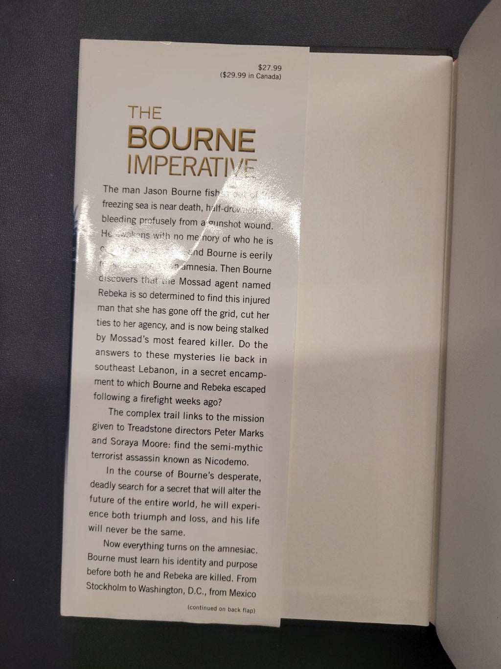 A glossy paper insert card for 'The Bourne Imperative' movie, featuring a white background with gold and black text, photographed inside a dark cardboard box.