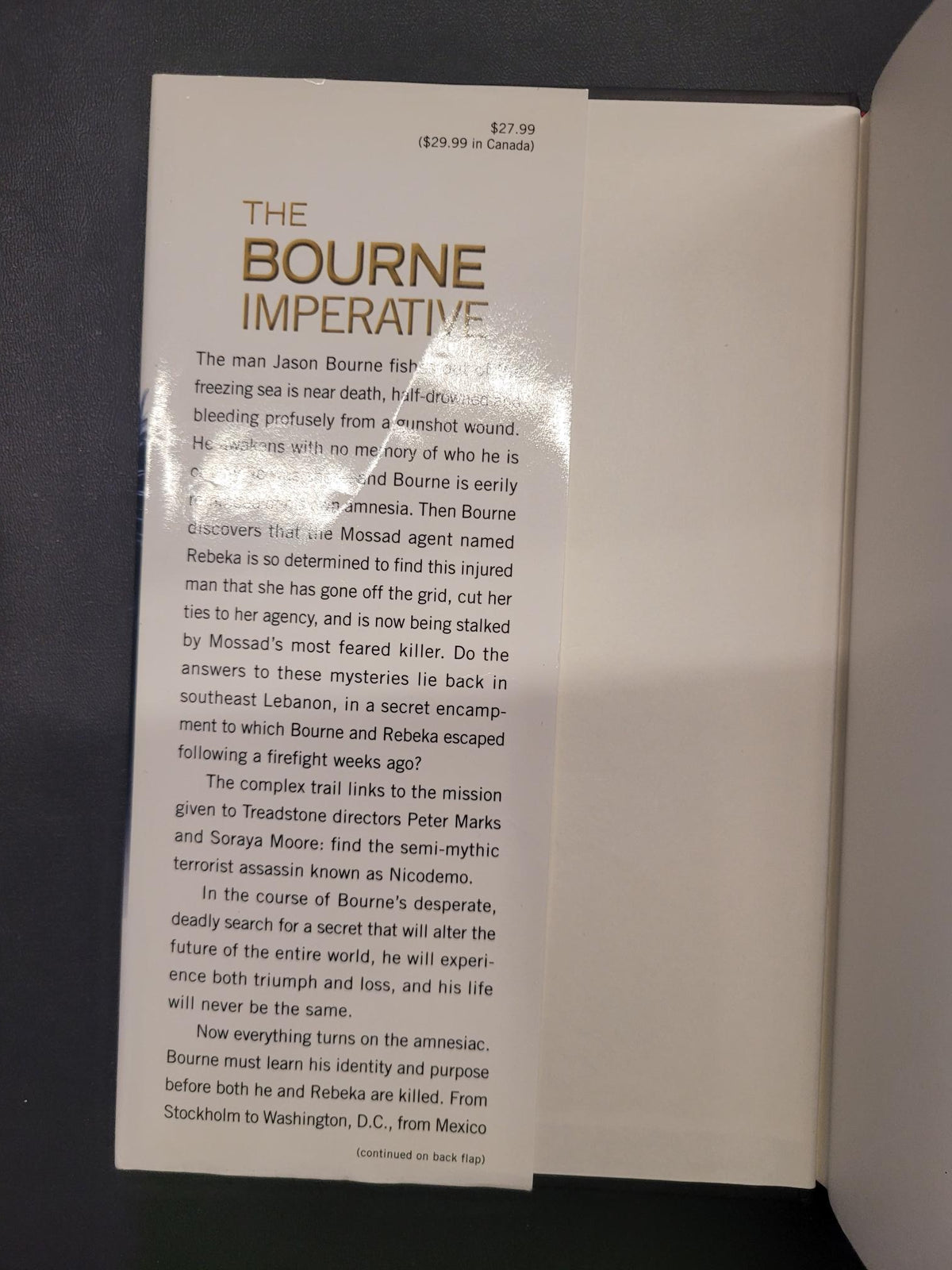 A glossy paper insert card for 'The Bourne Imperative' movie, featuring a white background with gold and black text, photographed inside a dark cardboard box.