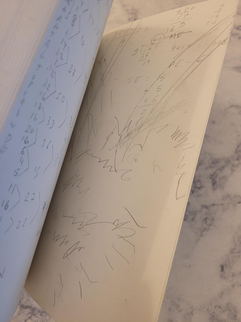 A close-up of a cream-colored notebook or journal with handwritten equations and sketches on the pages, resting on a light marble surface.