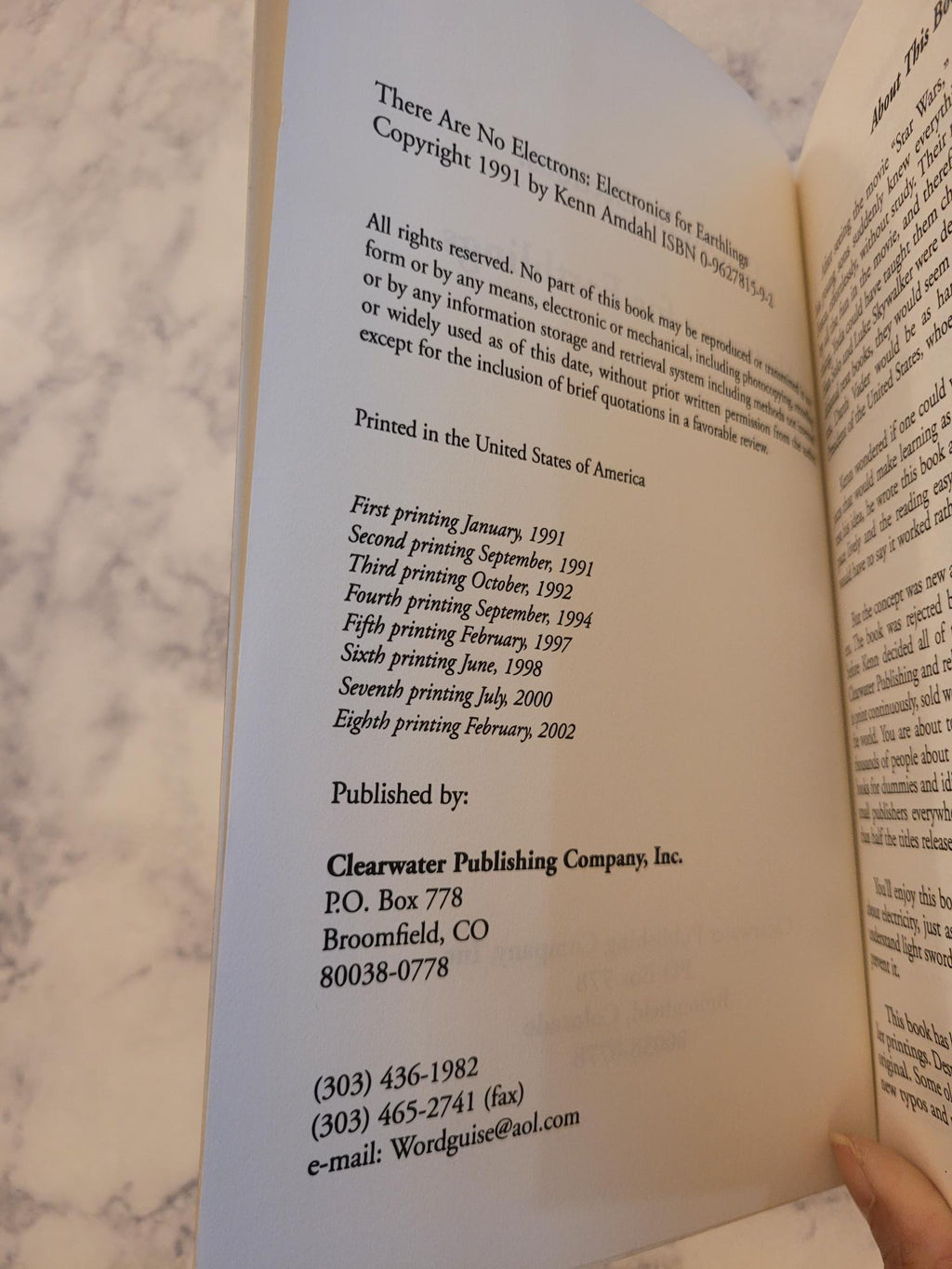 An open book showing printed text and copyright information, photographed against a light marble surface. A hand is visible holding the left edge of the book.