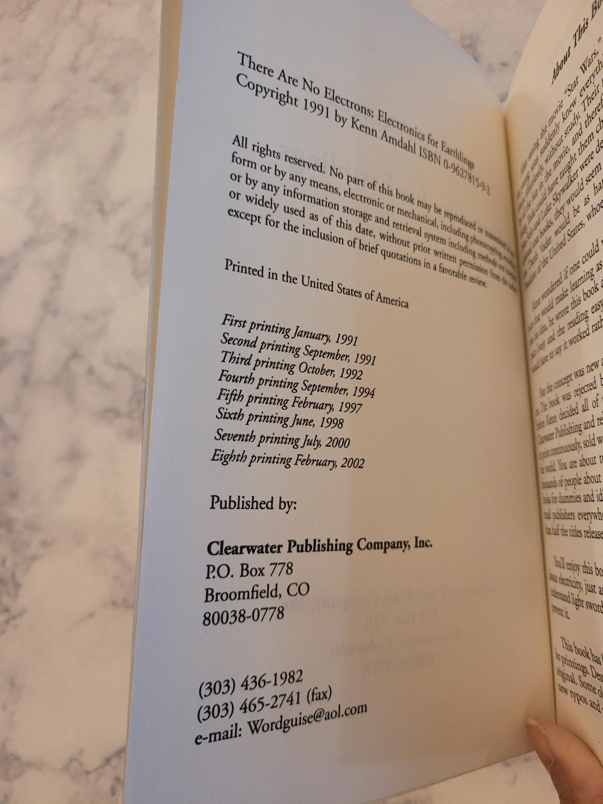 An open book showing printed text and copyright information, photographed against a light marble surface. A hand is visible holding the left edge of the book.