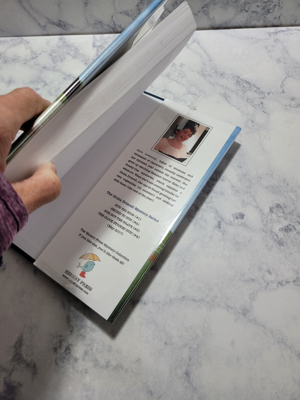 A person's hand and arm are visible as they open a printed booklet or book on a white and gray marble surface. The booklet pages are turned open, showing printed text and a small inset photo.